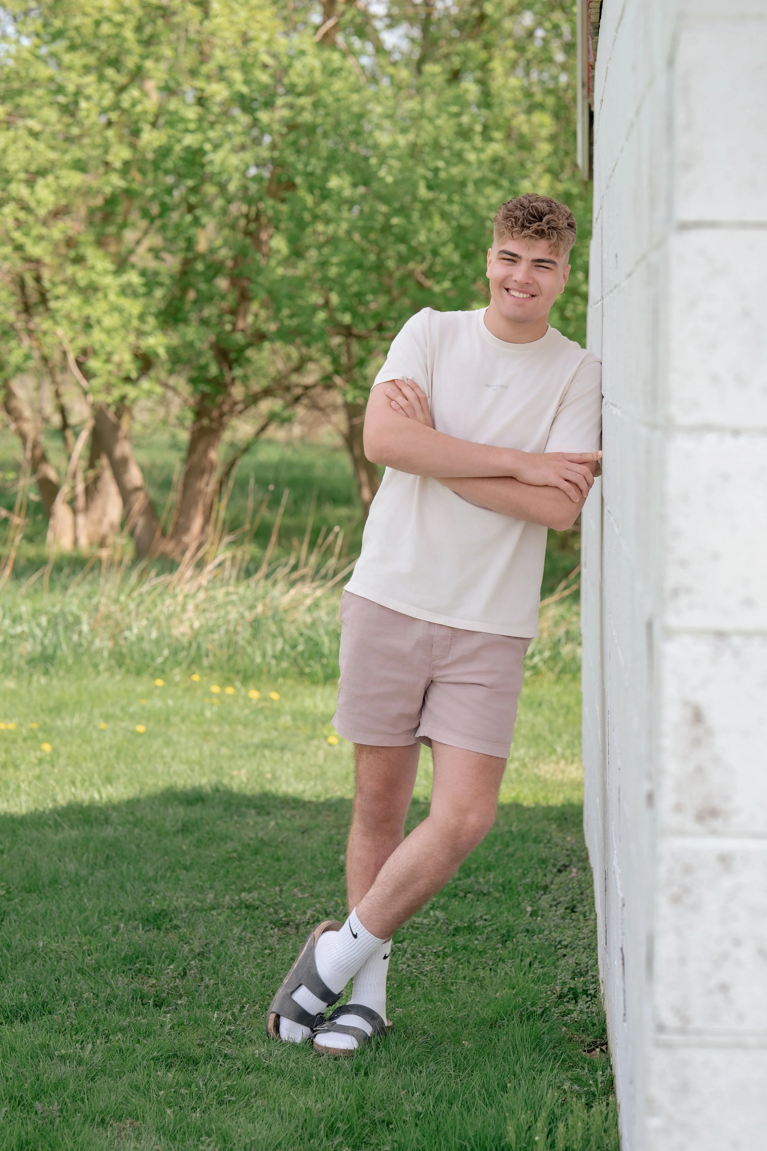 Young man leaning against a white wall outside, smiling, wearing a beige t-shirt, pink shorts, white socks, and sandals, with green trees and grass in the background.