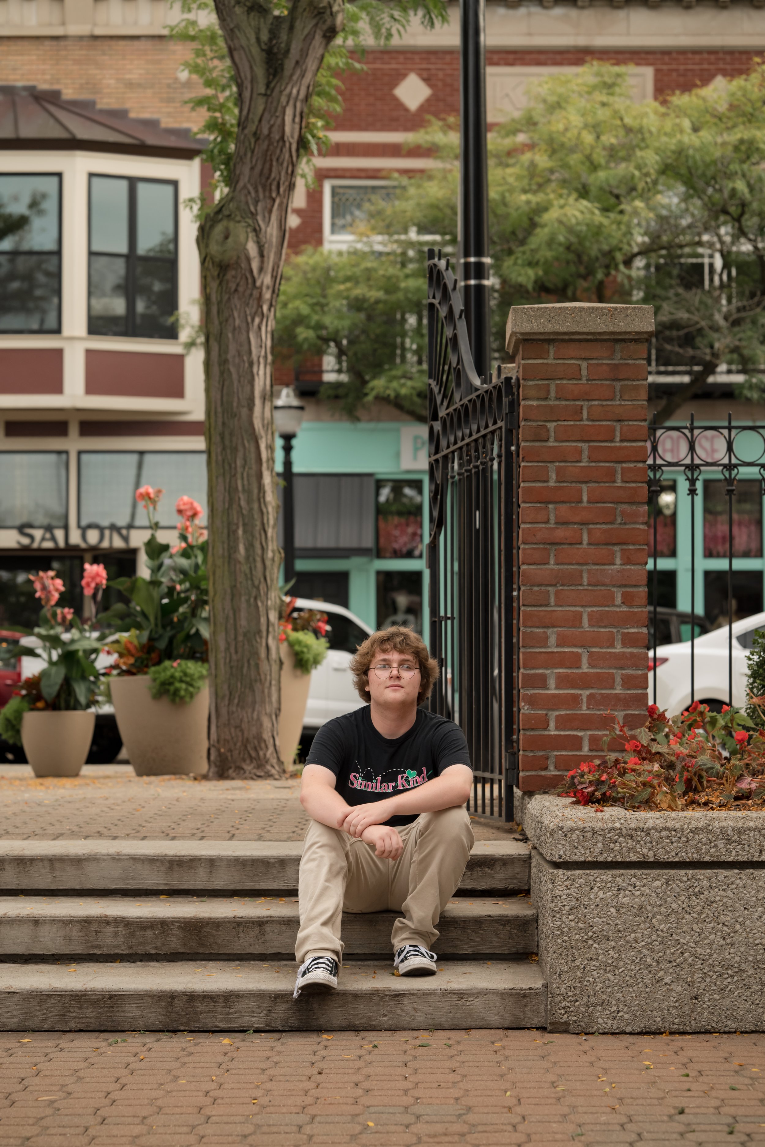 A young man with curly hair and glasses sitting on outdoor steps in front of a black wrought iron fence. There are potted flowers nearby, and trees, buildings, and parked cars in the background.