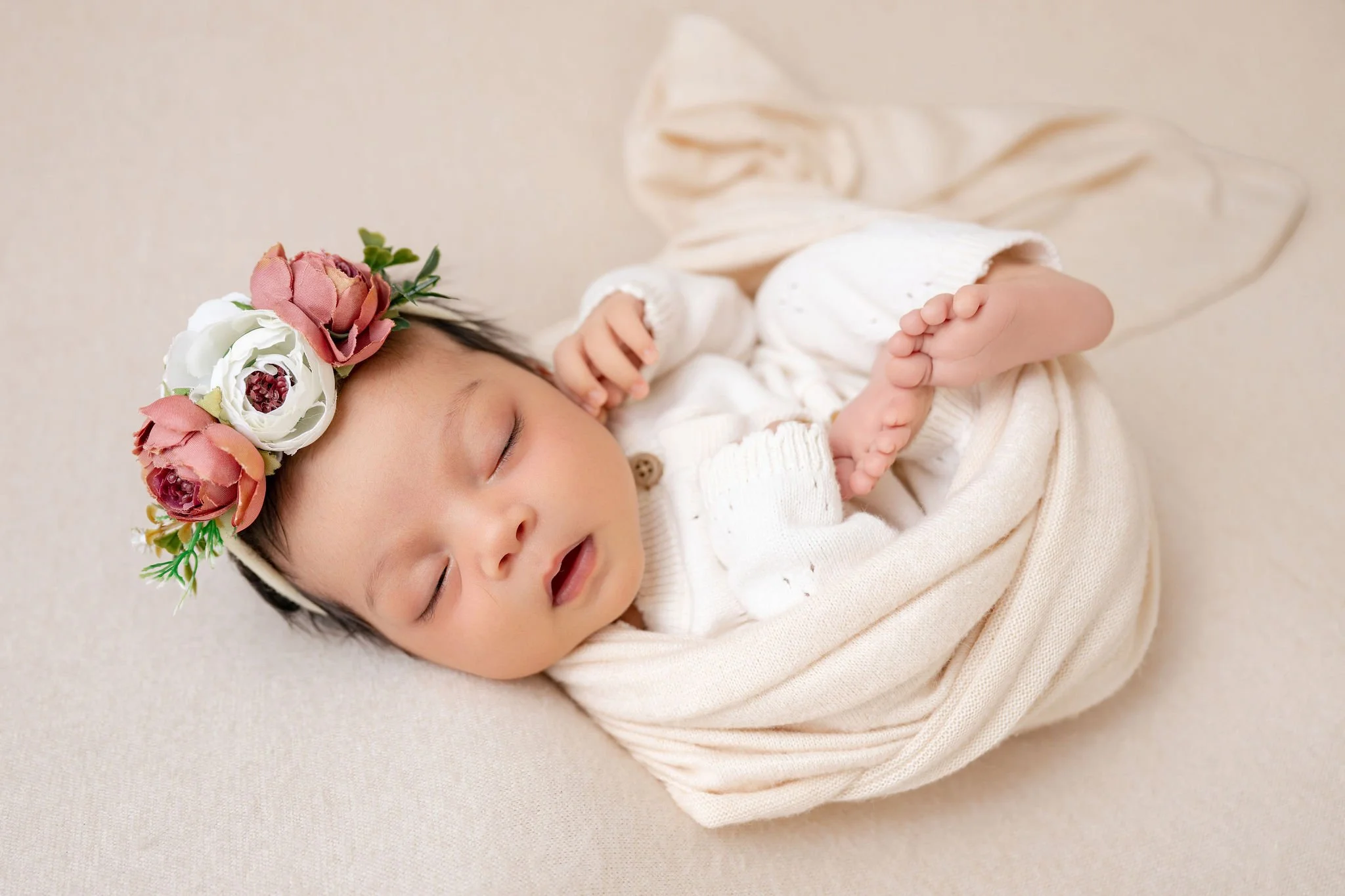 A sleeping baby girl wearing a flower headband lying on a soft beige surface.