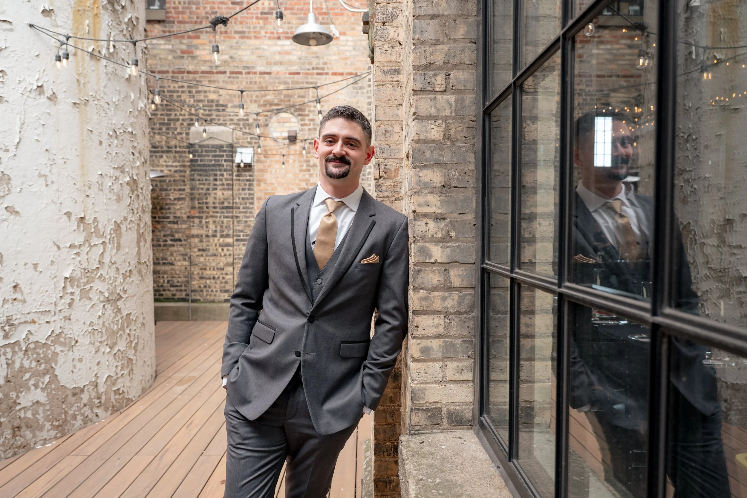 Groom leaning agains a brick wall near a window reflection and a dilapidated brick wall in the background