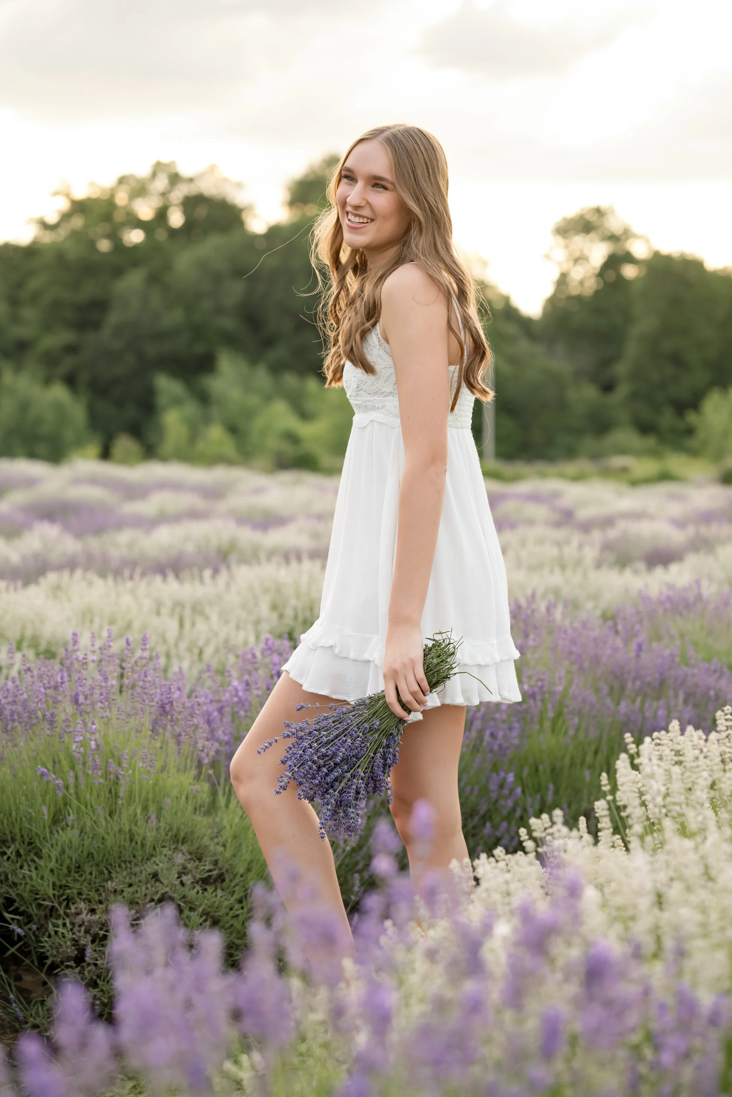 A young woman in a white dress standing in a lavender field holding a bouquet of lavender flowers, smiling and looking to the side during sunset.
