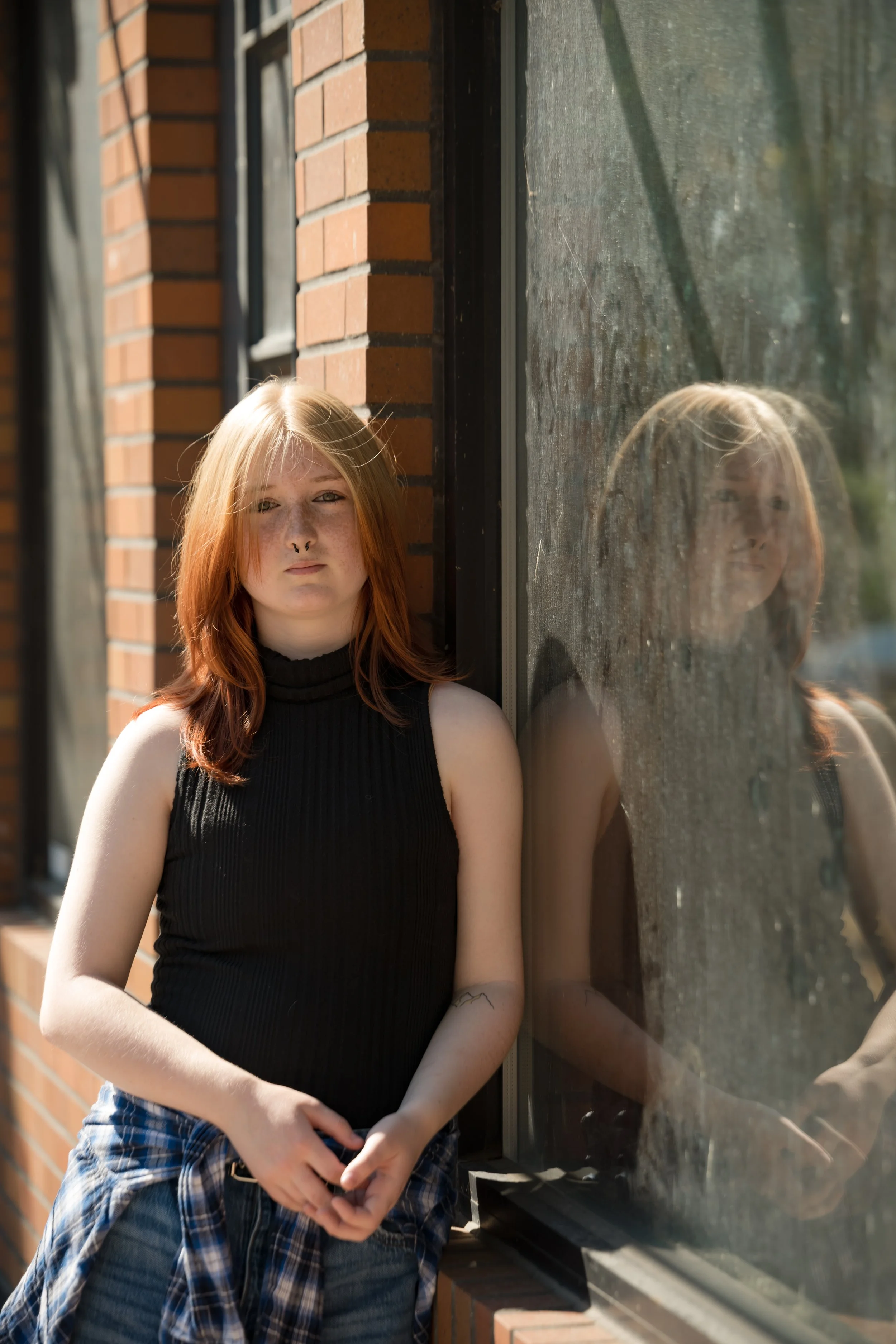 A young woman with red hair and freckles standing outdoors during daytime, leaning against a brick building, with her reflection visible in a nearby glass window. She wears a black sleeveless top and has a plaid shirt tied around her waist.