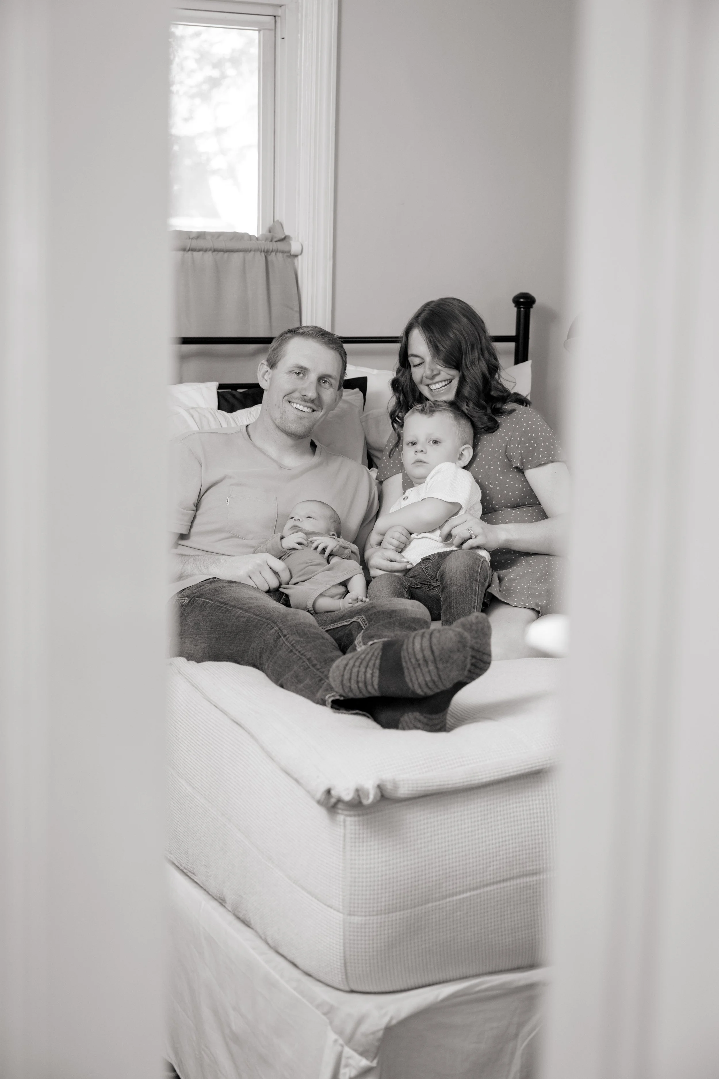 A black-and-white photo of a family sitting on a bed, framed through a slightly open door. The family consists of a man, a woman, and two young children, all smiling and looking at the camera. The room has a window with light curtains in the backgrou