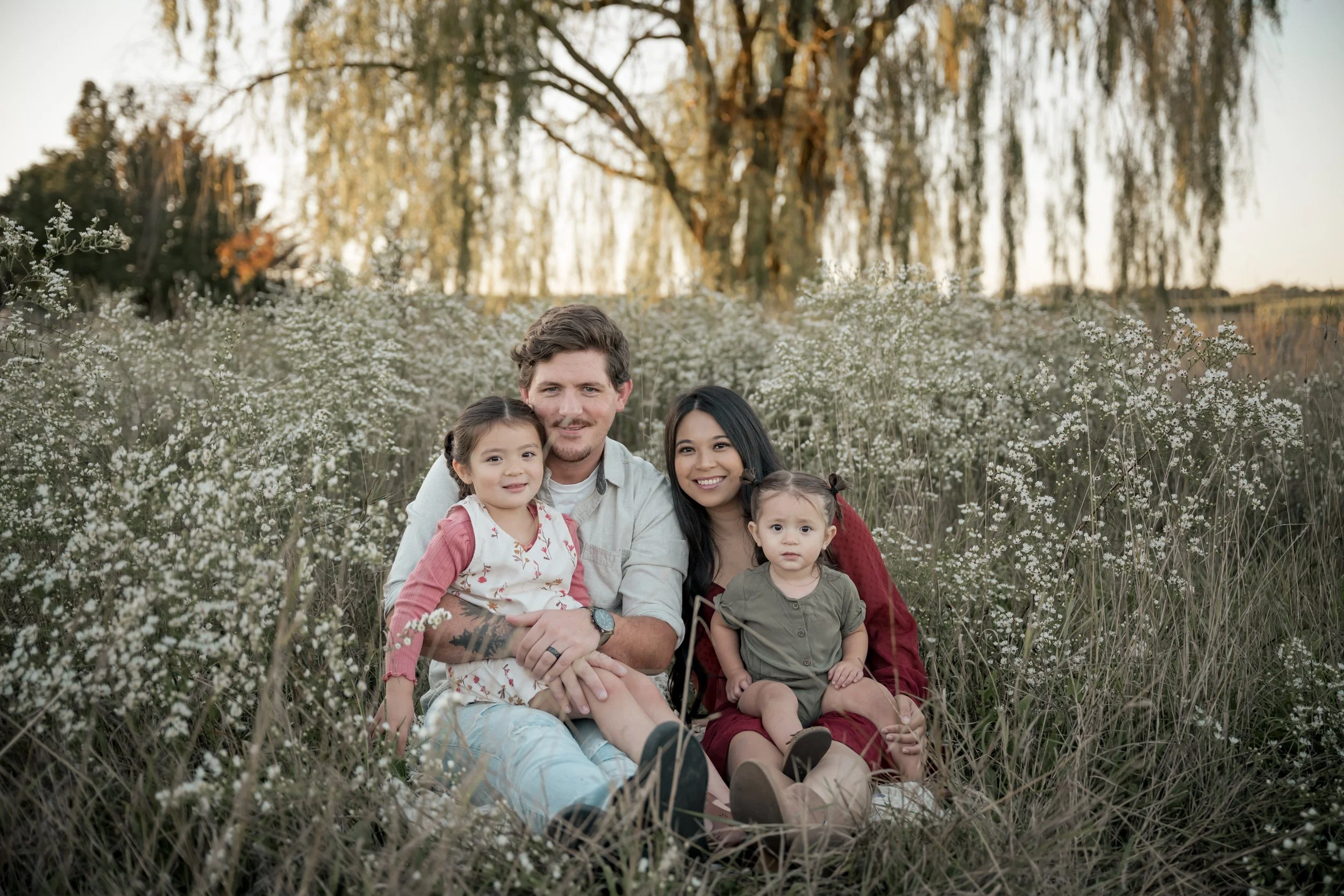 A family sitting in a field of white flowers with a willow tree in the background West Michigan Family Photography