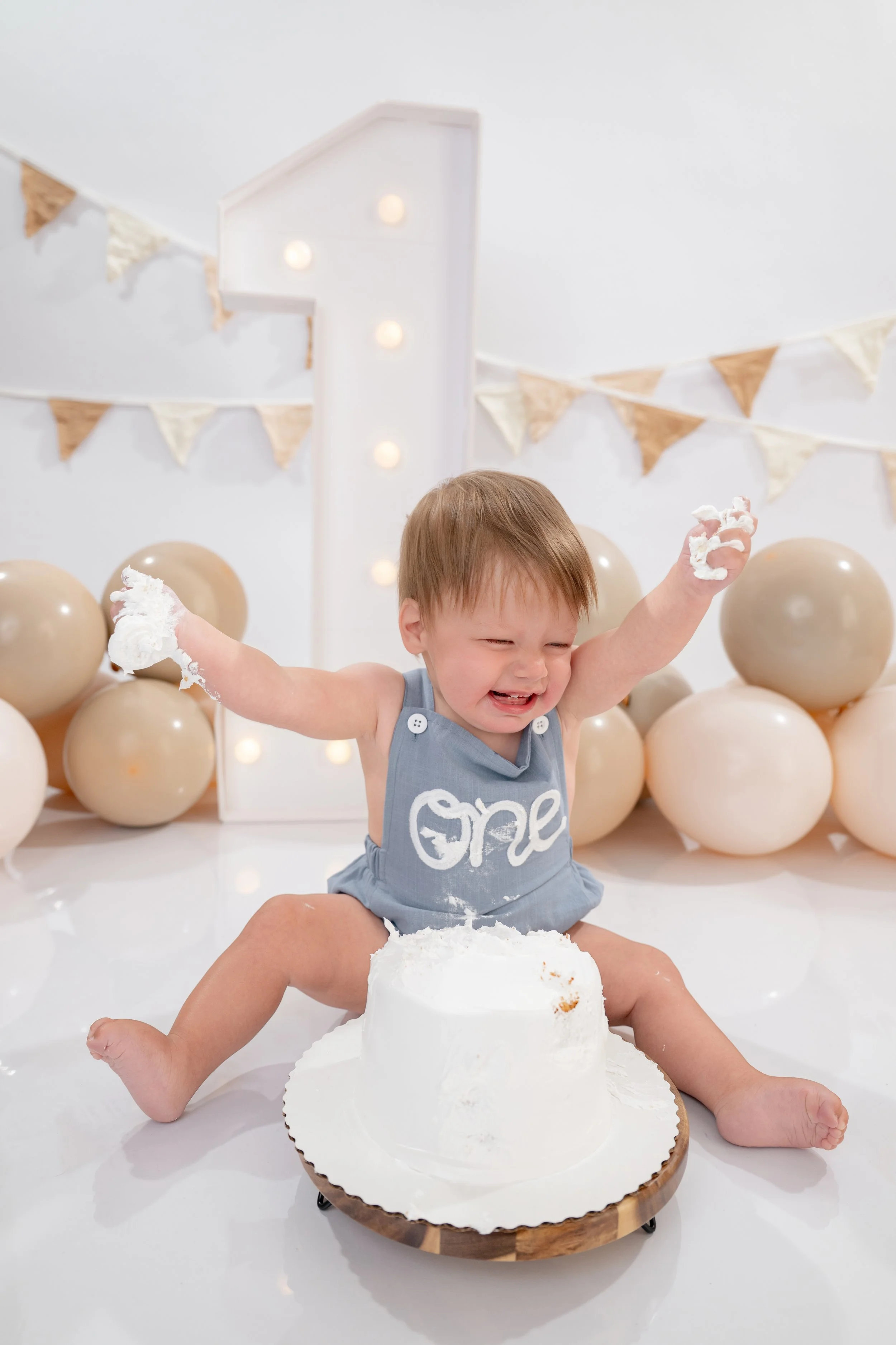 A young child with brown hair and a gray dress is playing with a white birthday cake on a round wooden tray. The child's arms and face are covered in white frosting, and they are smiling with eyes closed. The background has beige balloons, bunting, a