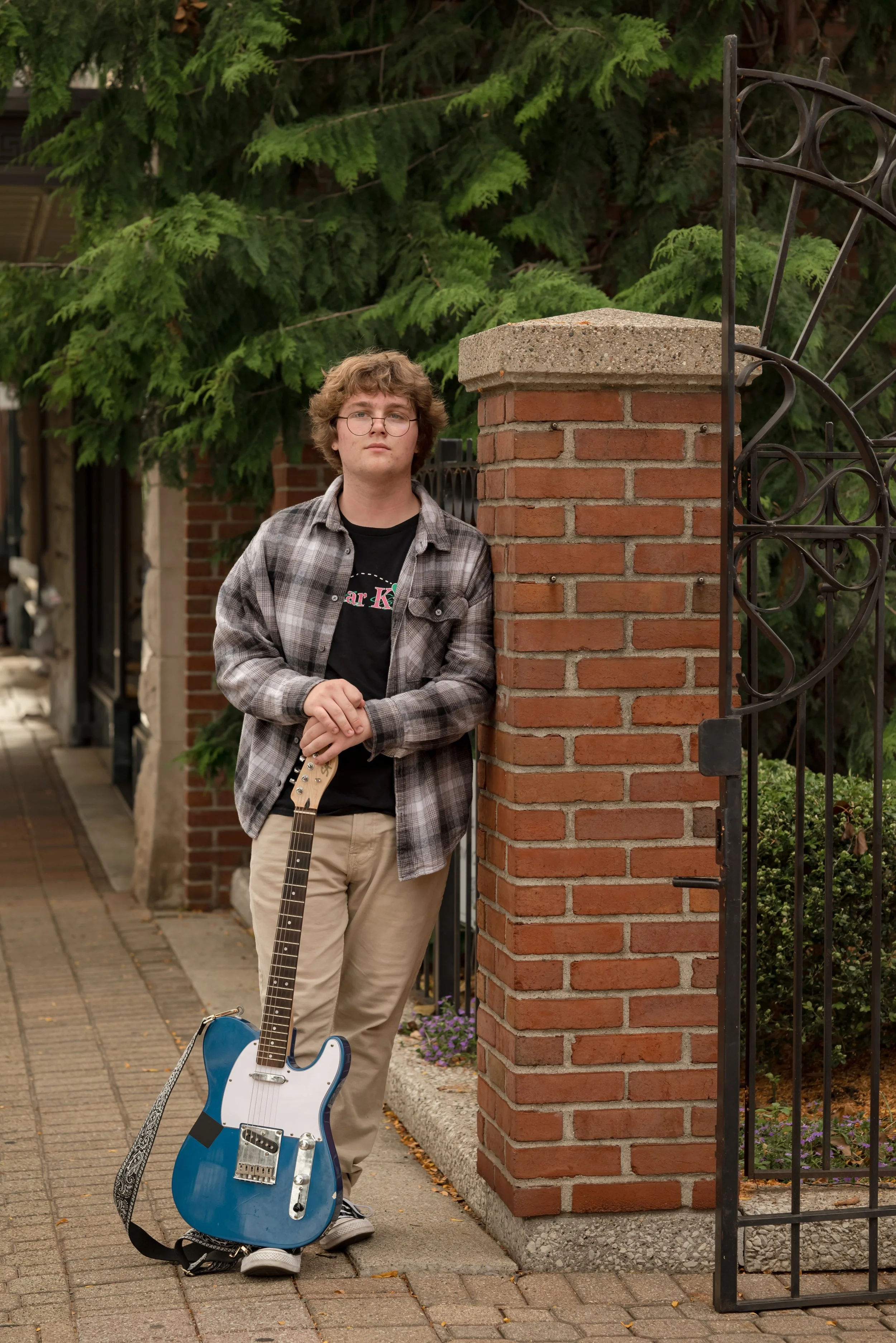 A young man with curly hair, glasses, and a black T-shirt under a plaid shirt, leaning against a brick pillar next to a black wrought iron gate. He is holding a blue electric guitar and has sneakers on. The background includes green trees and a sidew