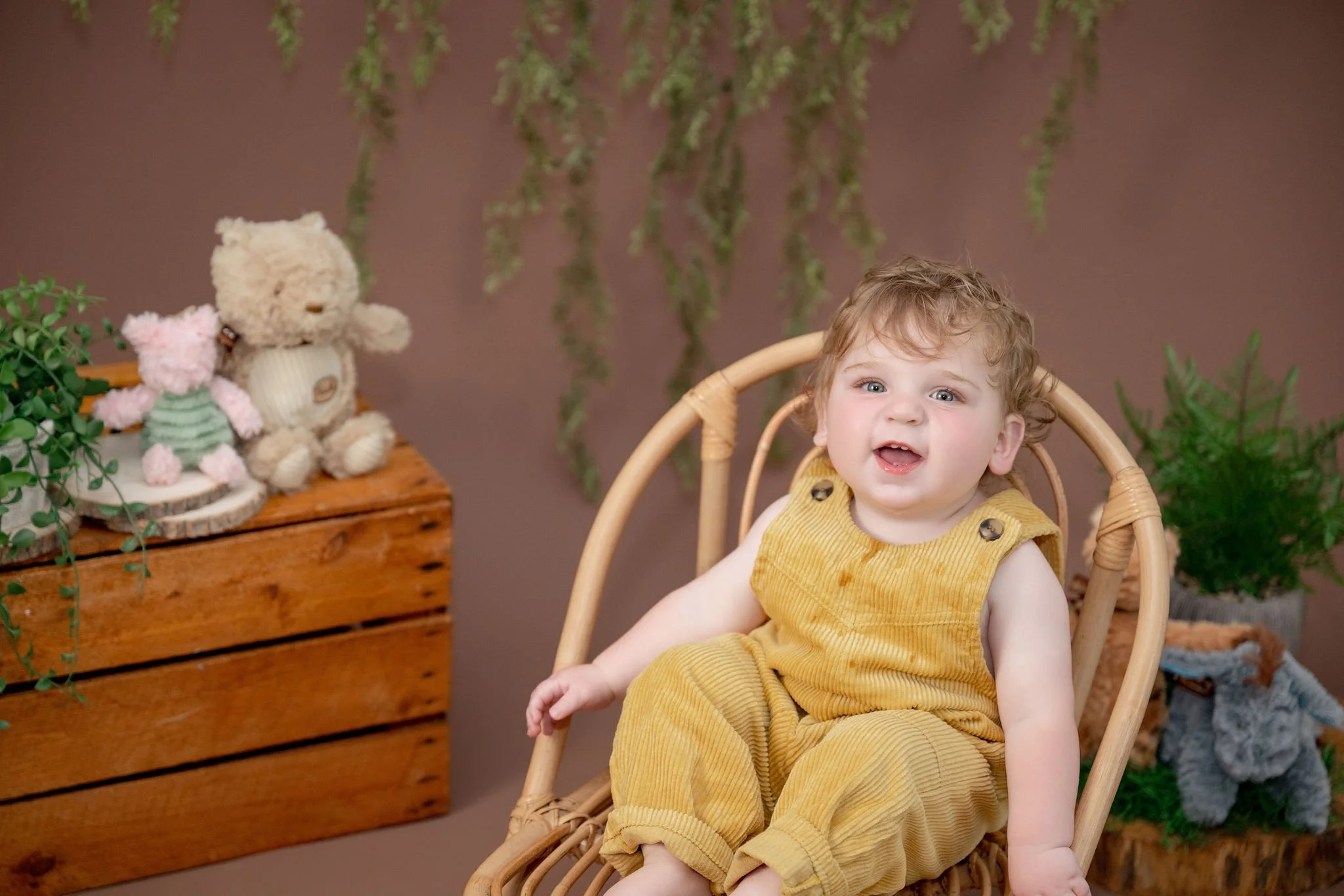 A young child with curly hair and blue eyes is sitting in a wicker chair, wearing a yellow corduroy outfit. The child has a joyful expression, with mouth slightly open. Behind, there are green plants, a wooden chest with stuffed animals on top, and a