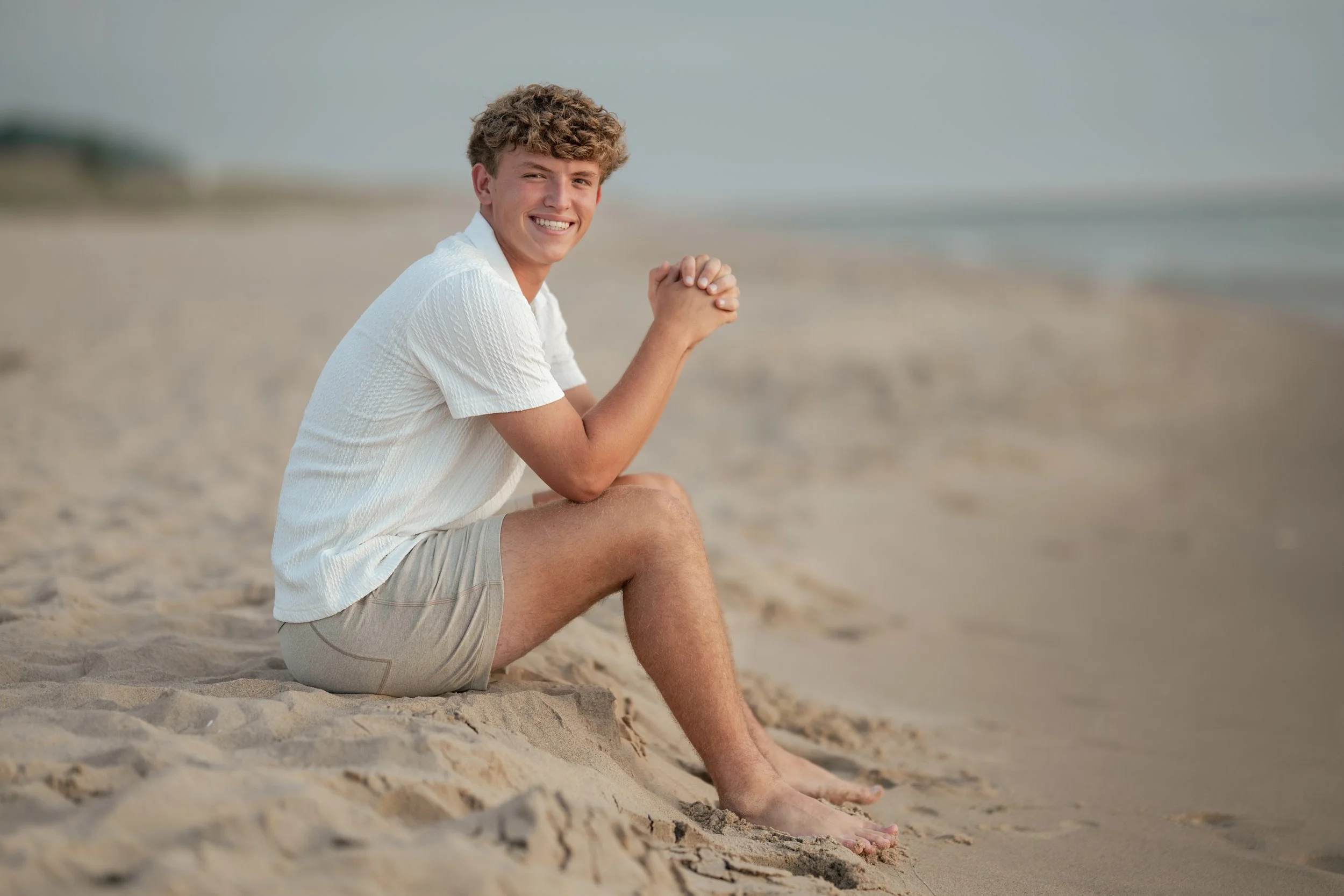 A young man with curly hair sitting on the sandy beach, smiling, wearing a white shirt and beige shorts, with the ocean in the background.