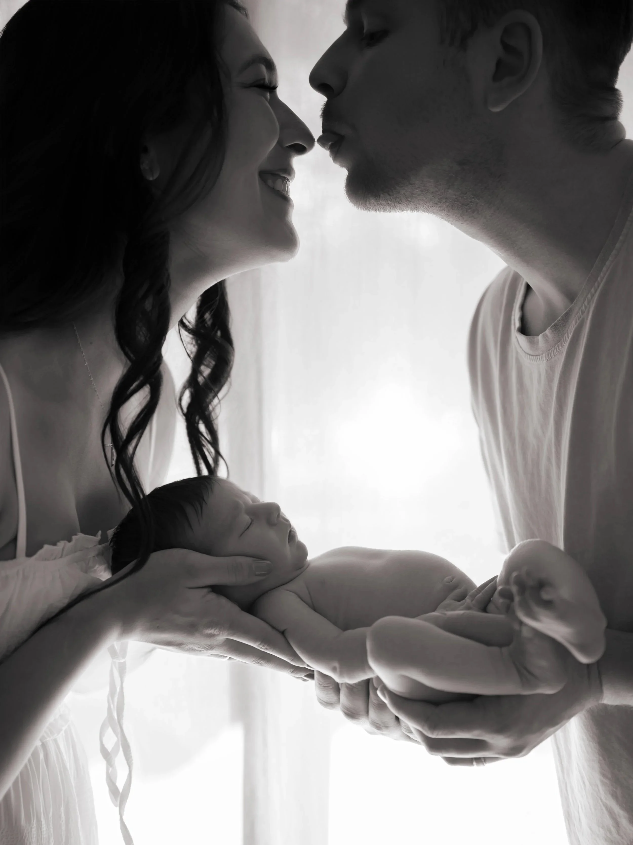 Black and white image of newborn baby posed in the parents hands with a window behind