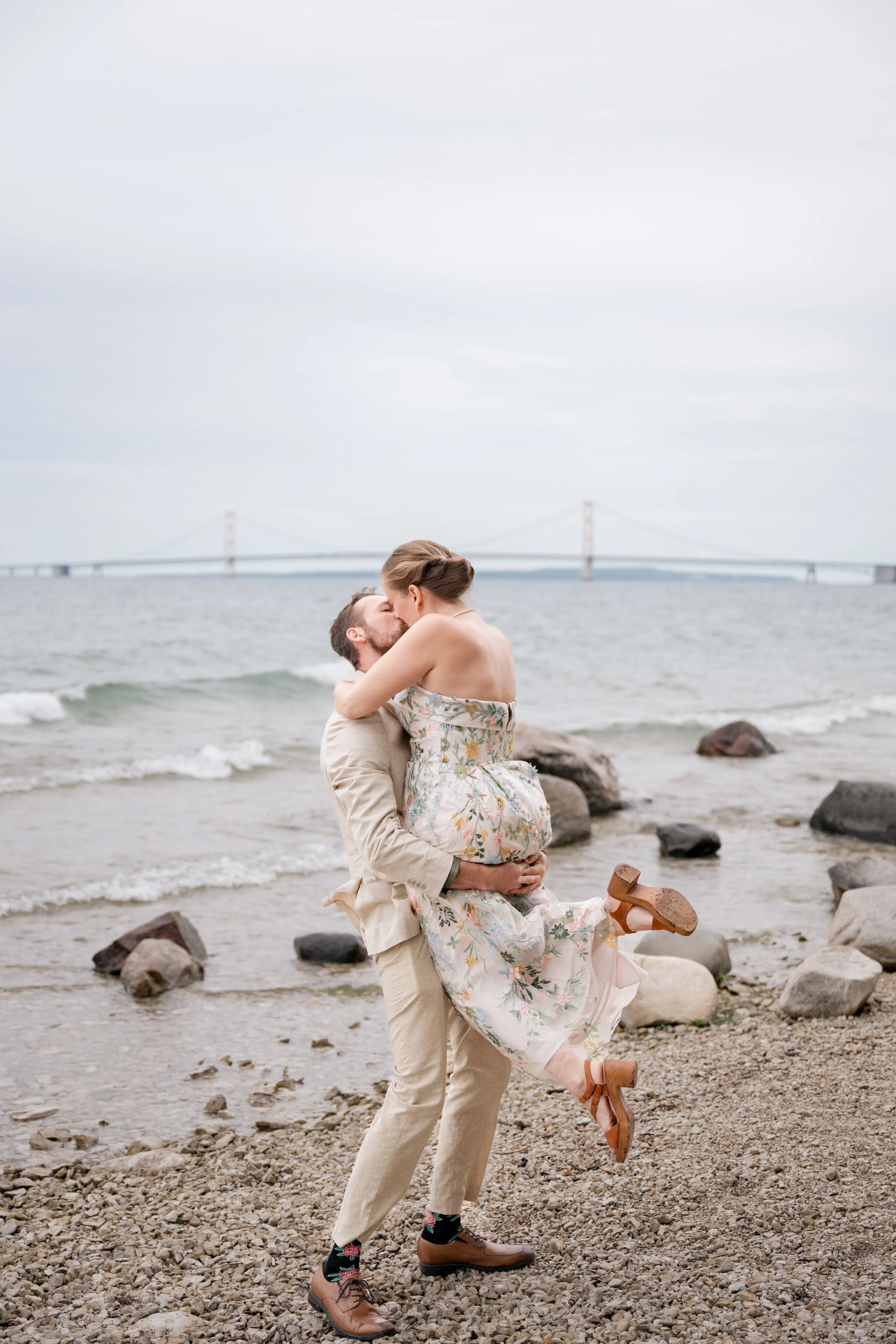 A man lifting a woman in a floral dress on a rocky beach with a bridge in the background.