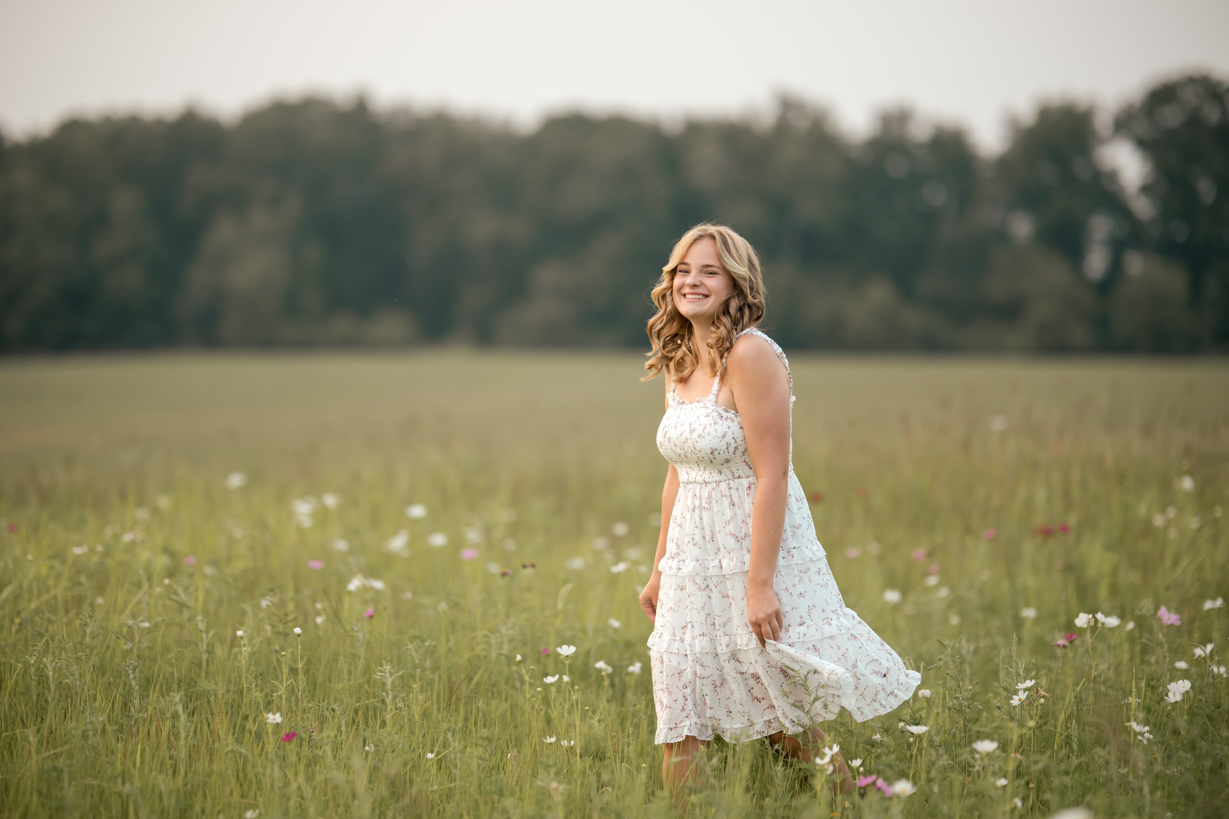 A young woman with wavy blonde hair wearing a white floral dress, standing in a field of wildflowers with a backdrop of trees and an overcast sky, smiling.