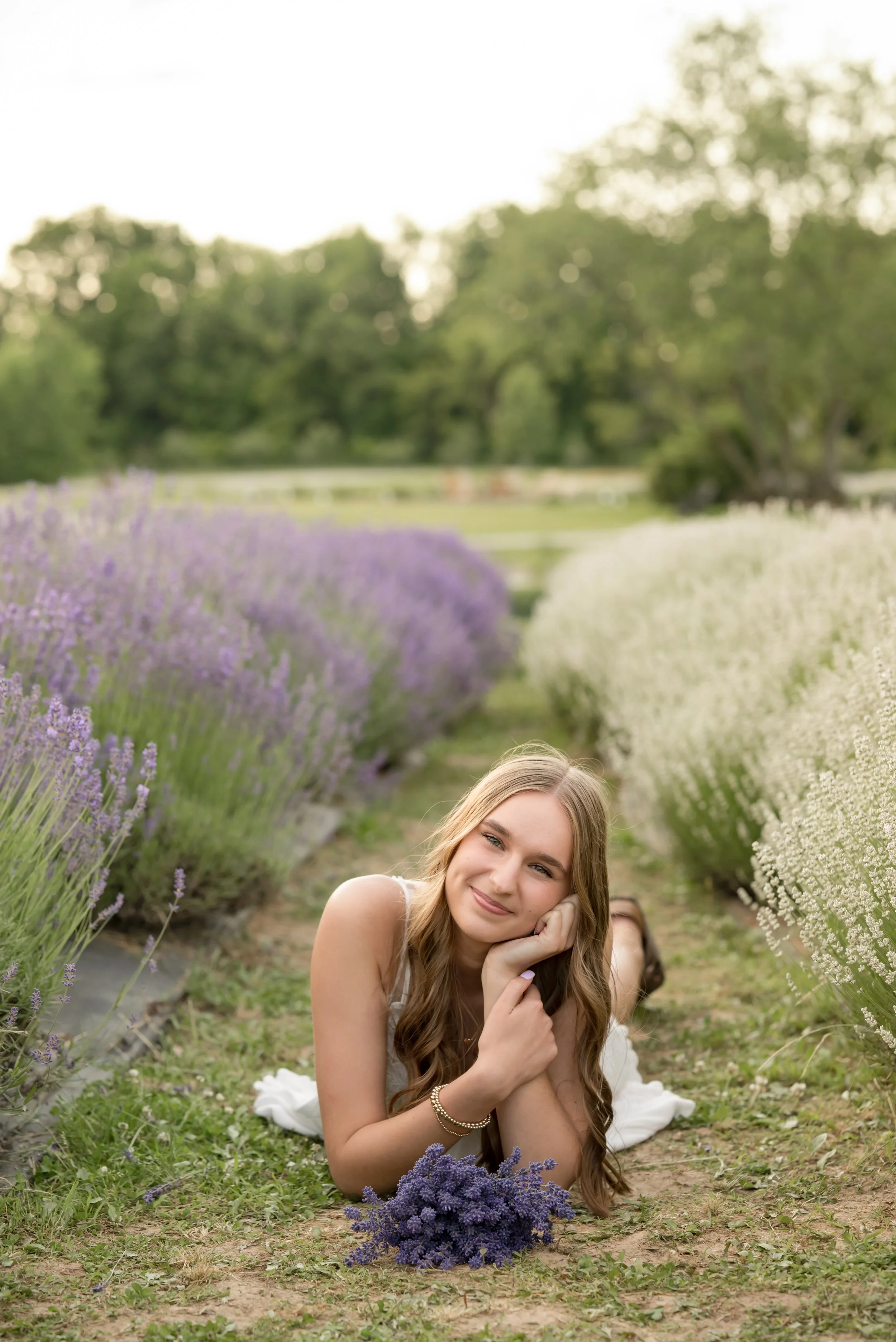 A young woman with long wavy brown hair lying on the ground in a lavender field, smiling peacefully, with her face resting on her hand, surrounded by purple and white lavender flowers and greenery, during daytime.