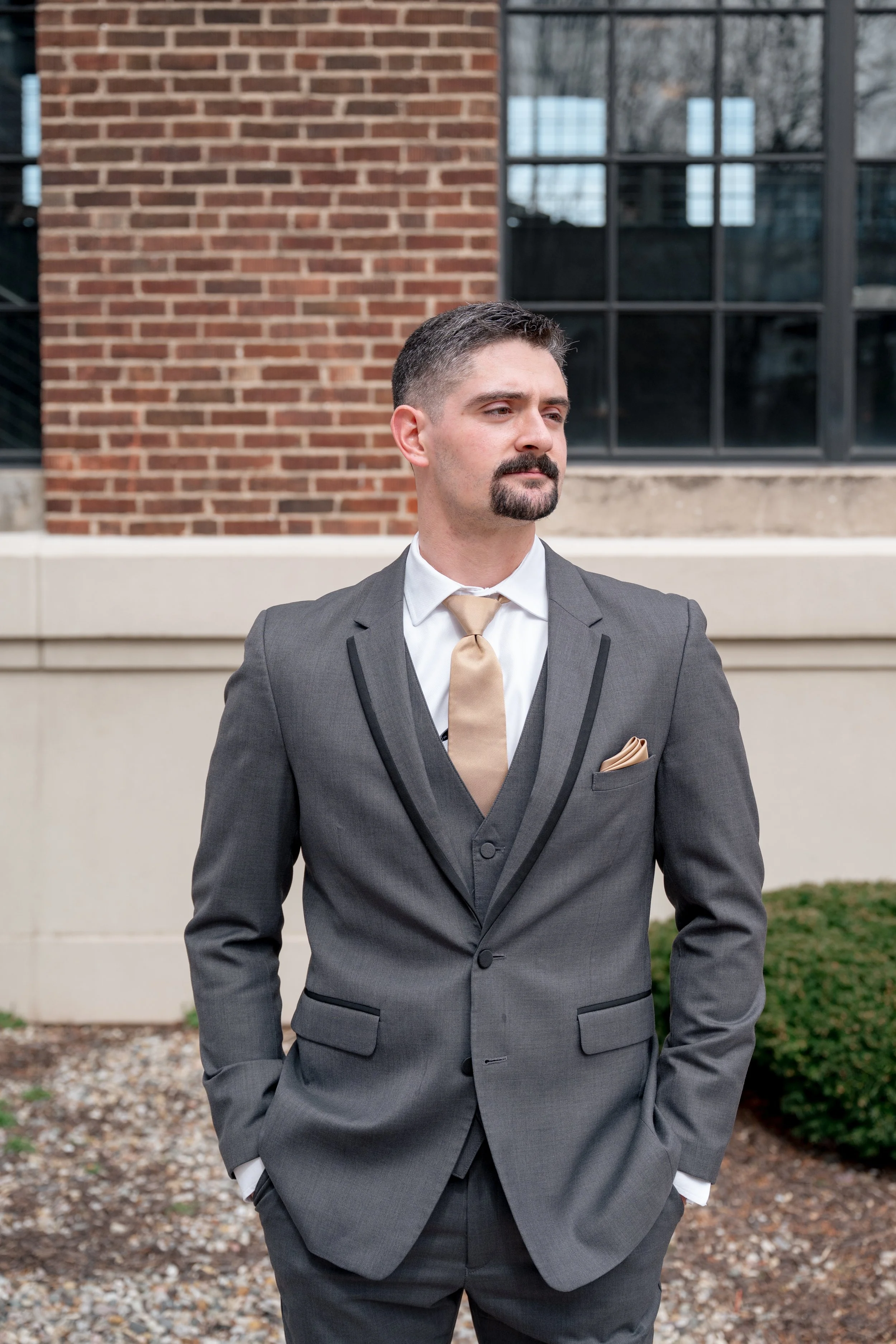 Groom standing in front of a brick building and windows. 