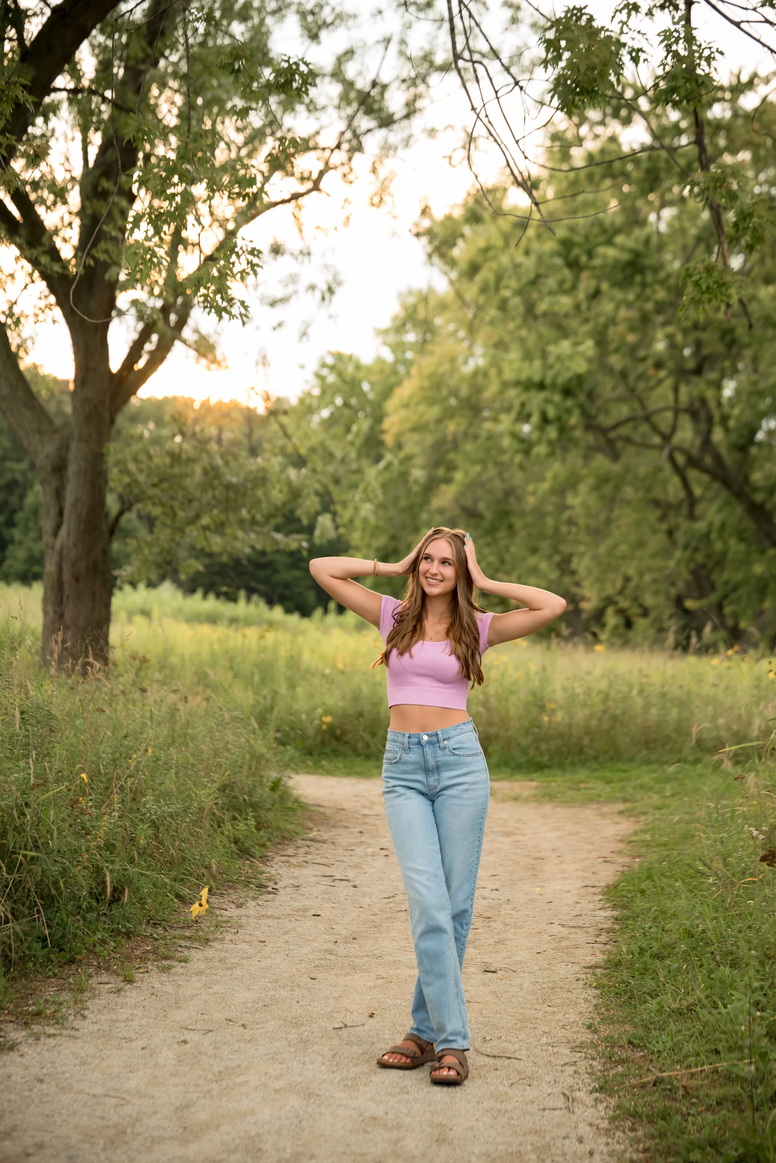 A young woman walking on a dirt trail through a green park during late afternoon or early evening, surrounded by trees and grass, wearing a pink crop top, blue jeans, and sandals, smiling and holding her hands behind her head.