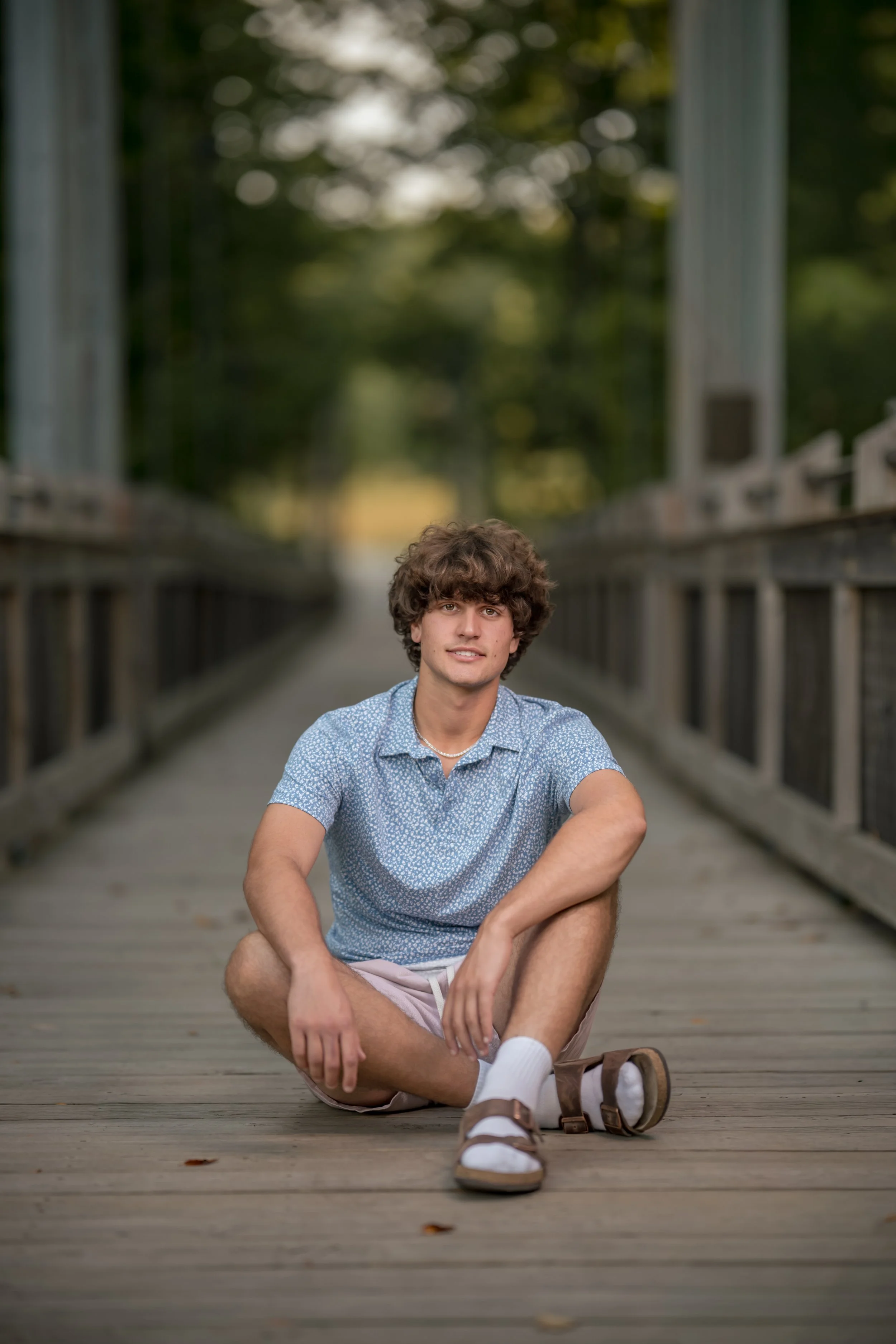A young man with curly brown hair and light skin sitting cross-legged on a wooden bridge, smiling at the camera, wearing a blue patterned shirt, beige shorts, white socks, and brown sandals, with trees and greenery in the background.