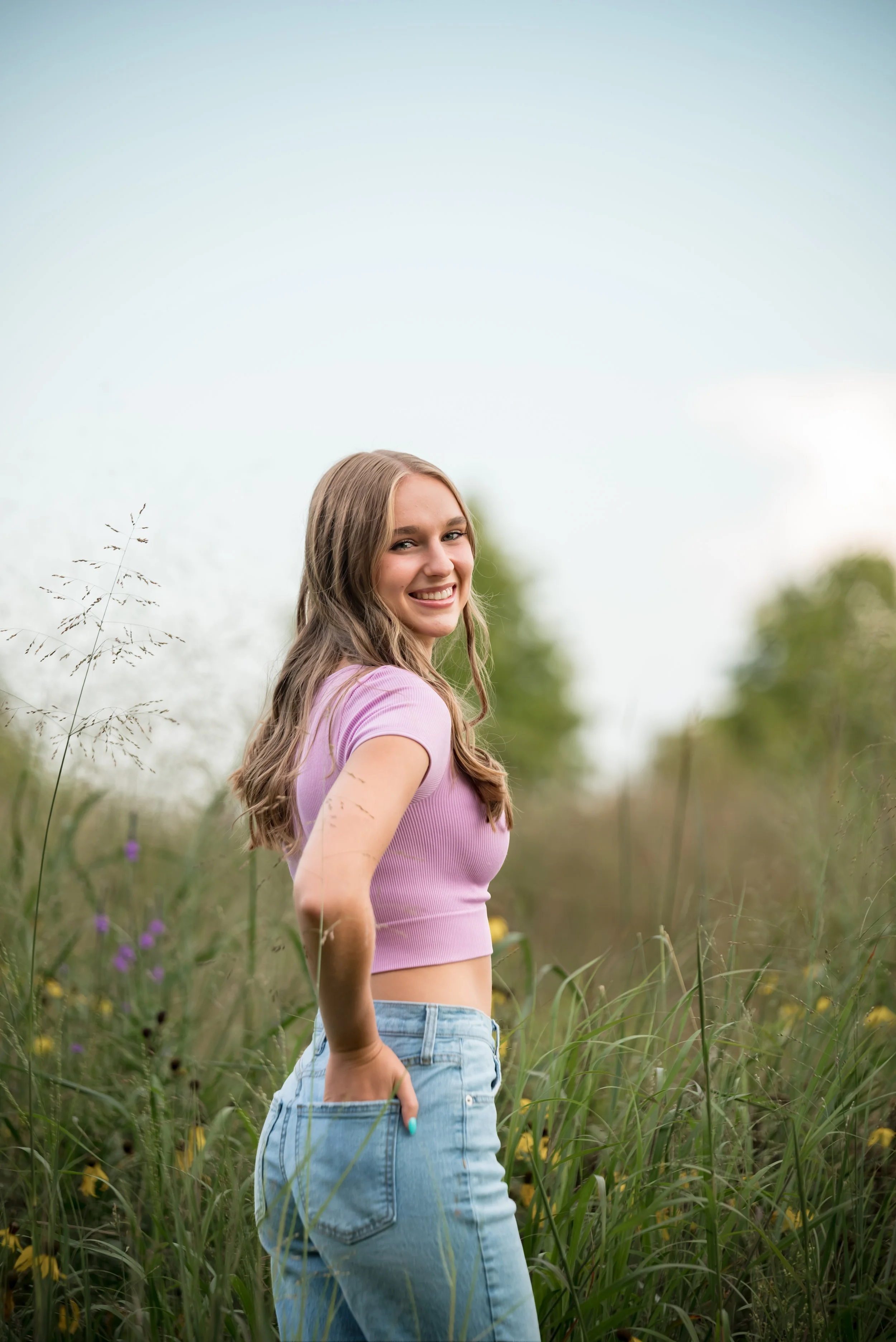 A young woman with long, light brown hair and a bright smile standing in a field of tall grass and wildflowers during daytime, wearing a light purple crop top and light blue jeans.