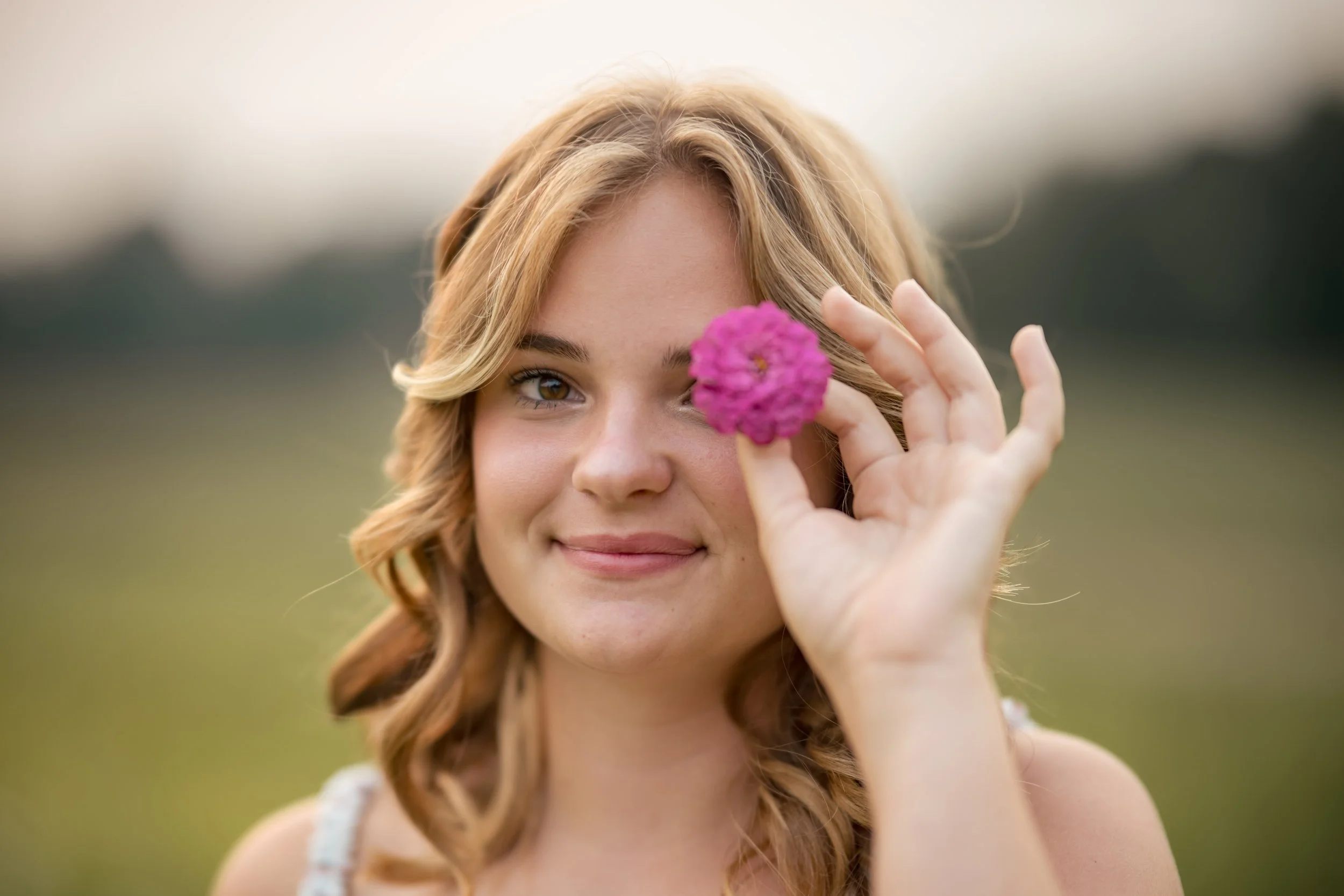 A young woman with blonde curly hair smiling outdoors while holding a pink flower in front of her eye.