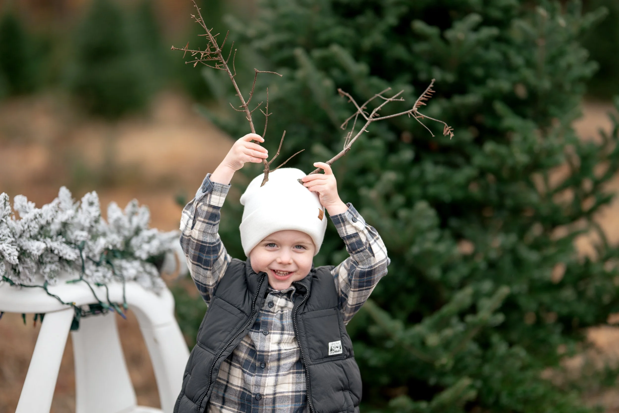 Young boy with blue eyes smiling and wearing a white knit cap, plaid shirt, and black vest, holding two brown tree branches above his head outdoors, with a snow-covered chair on the left and green bushes in the background.