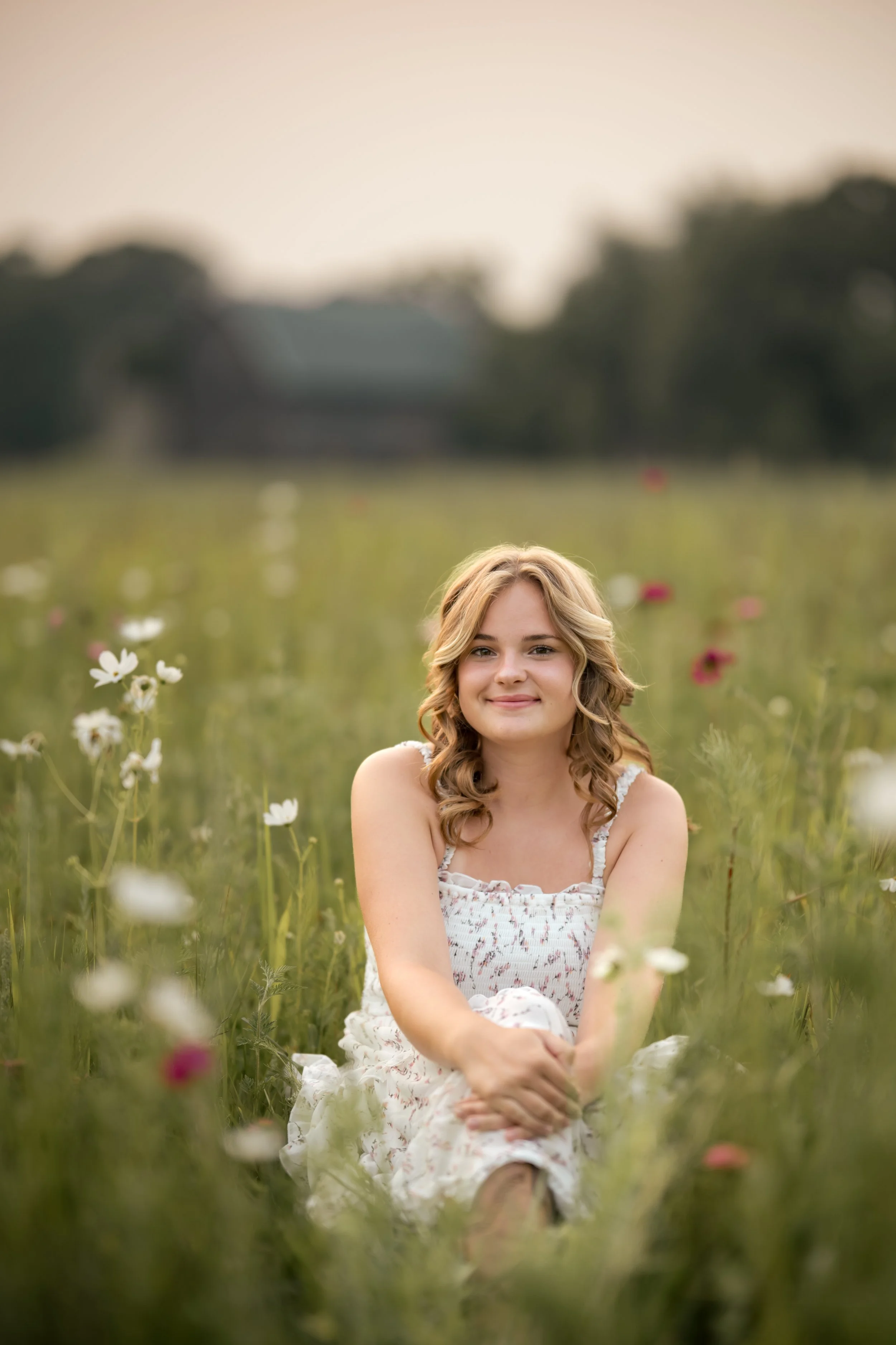A young woman sitting in a field of flowers with a gentle smile on her face, surrounded by blooming flowers and greenery, during sunset.