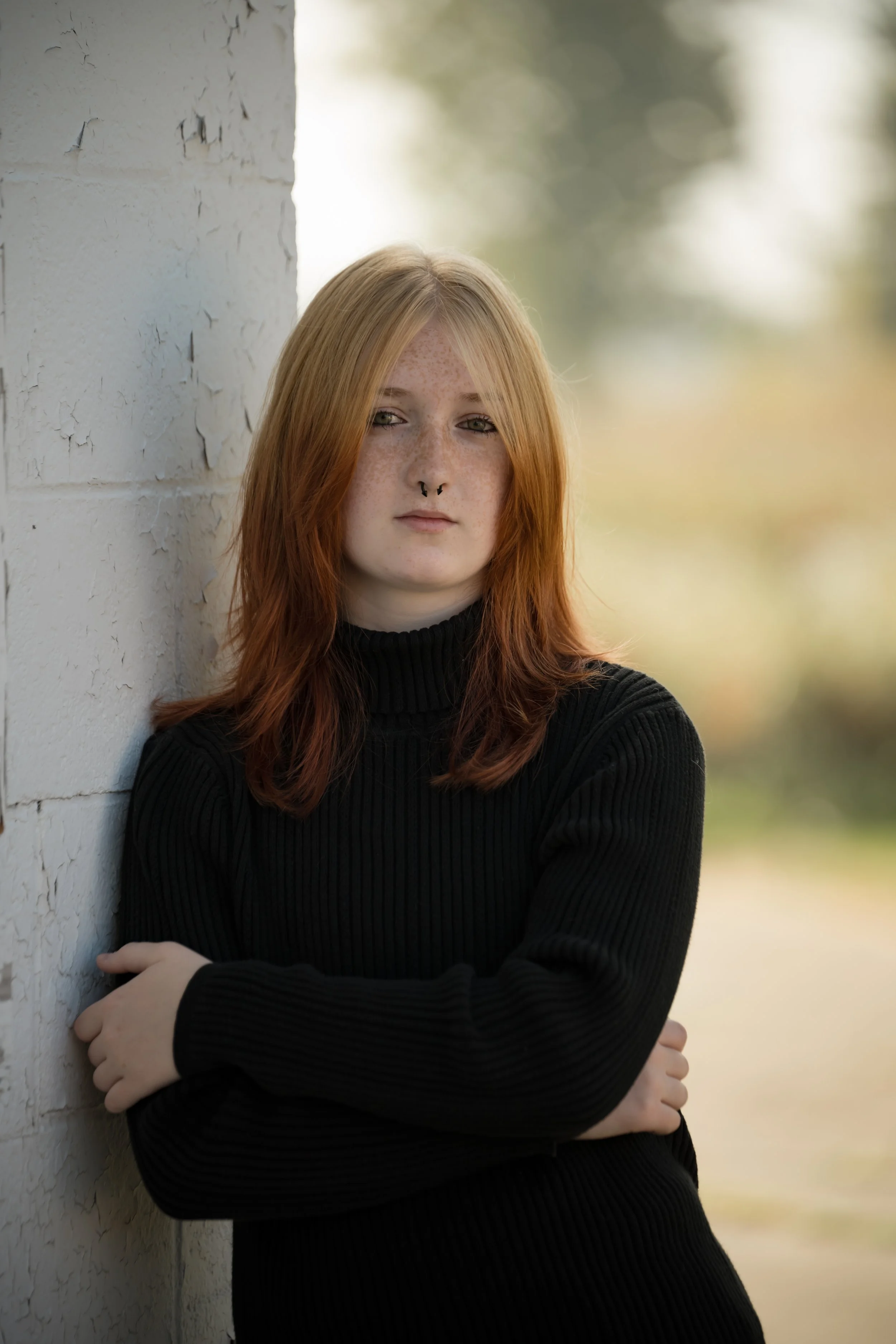 A young woman with red hair and freckles leaning against a textured white wall, outdoors with blurred greenery in the background.