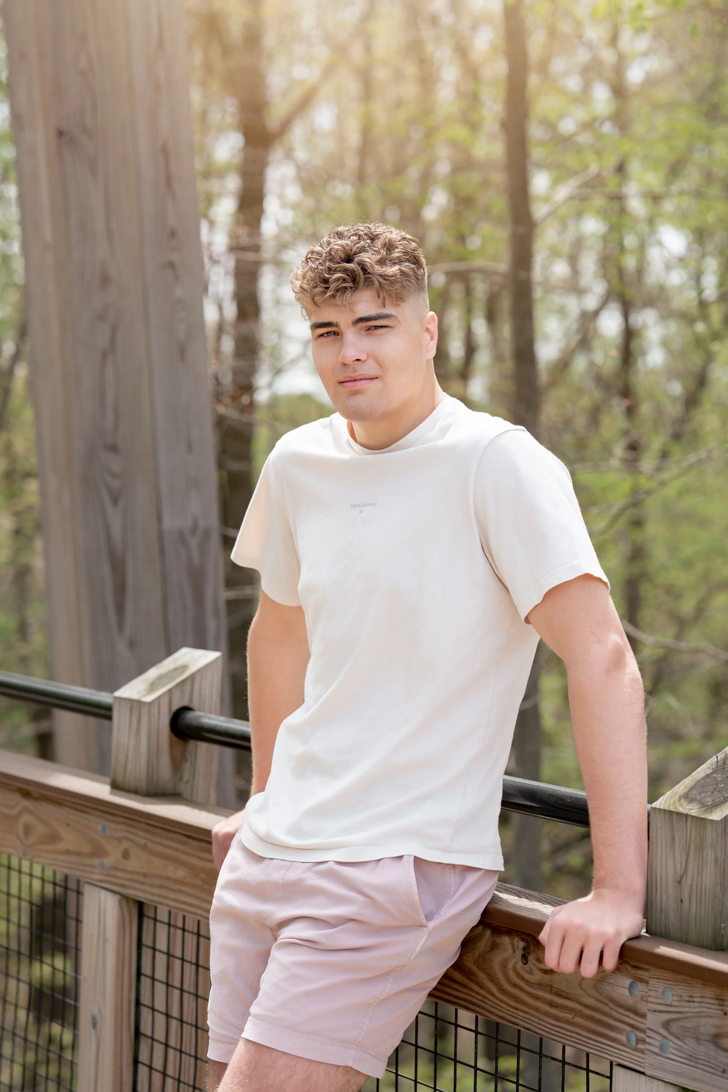 A young man with curly hair standing outdoors during daytime, leaning against a wooden railing on a platform surrounded by trees with green leaves.