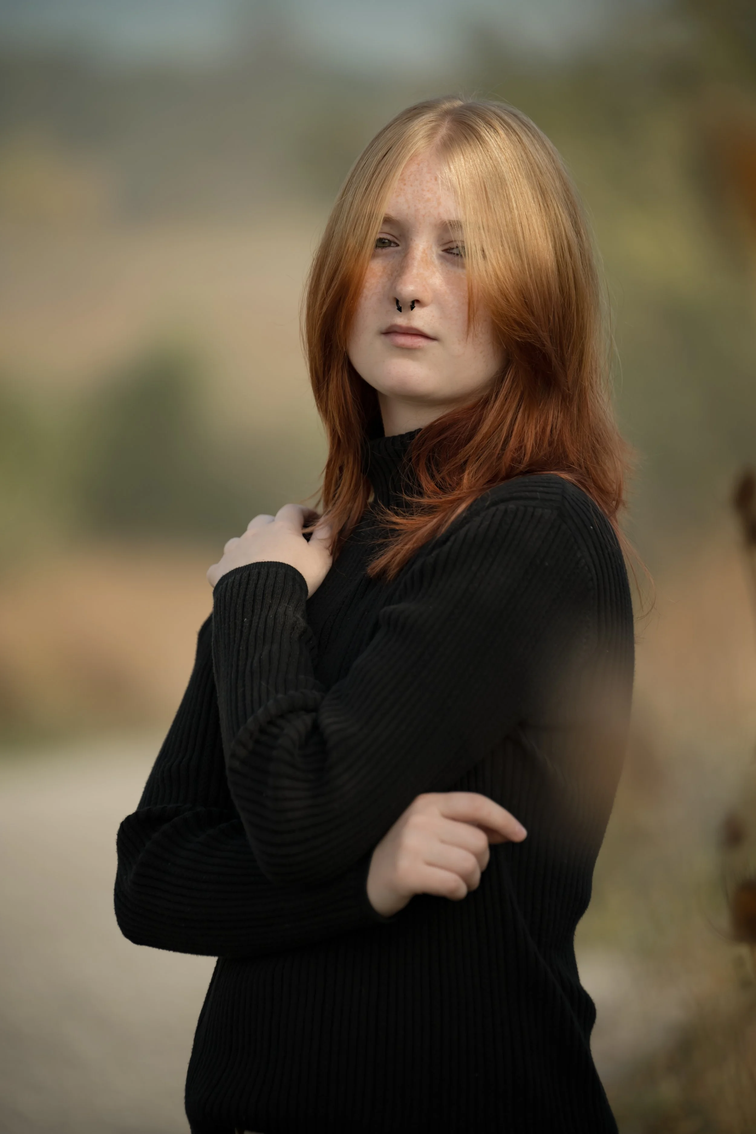 A young woman with red hair, freckles, and a septum piercing, wearing a black ribbed long-sleeve top, standing outdoors with a blurred natural background.