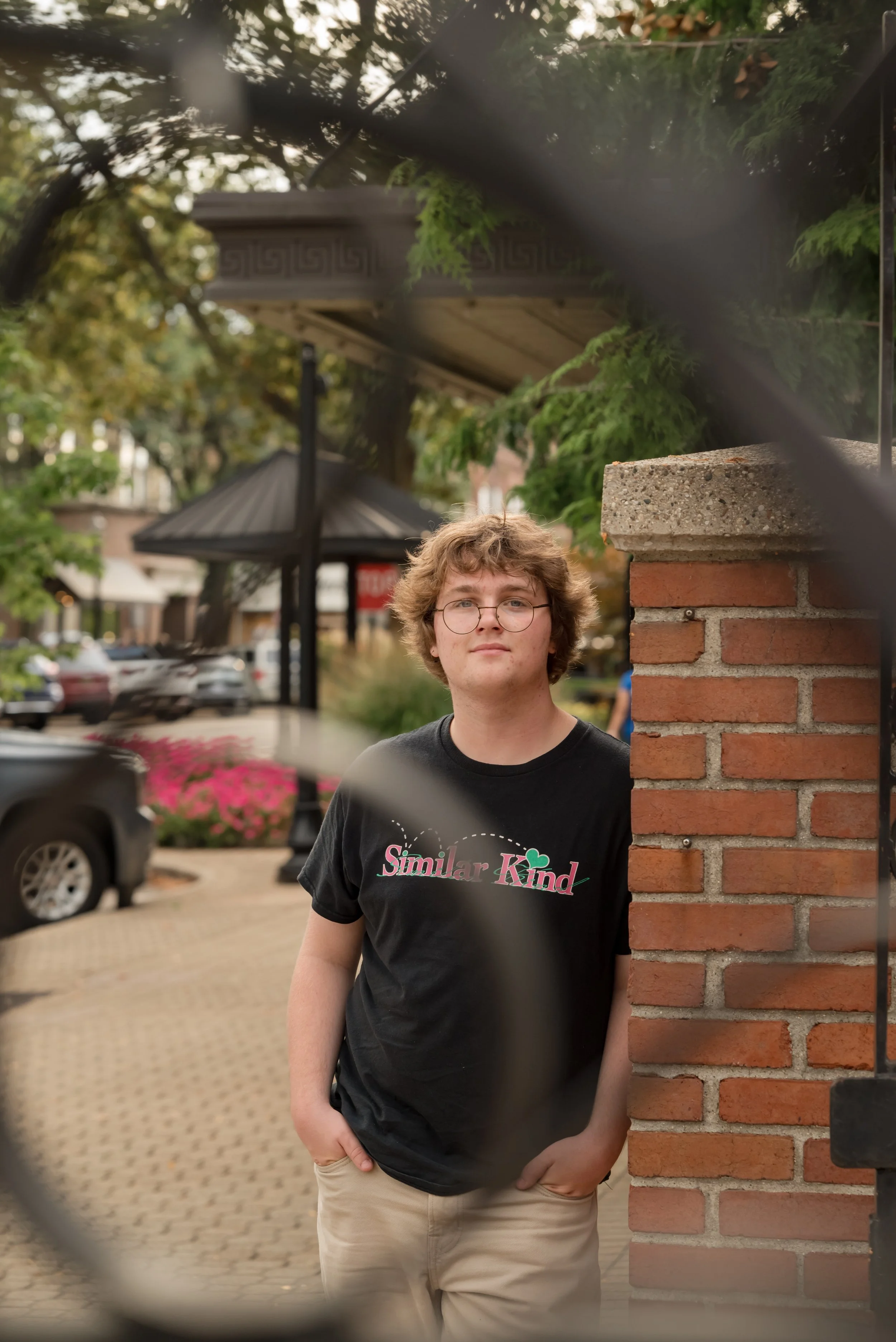 A young man with curly hair and glasses, wearing a black T-shirt with the words 'Similar Kind' and beige pants, standing outside near a brick wall and trees, with a landscaped sidewalk and parked cars in the background.