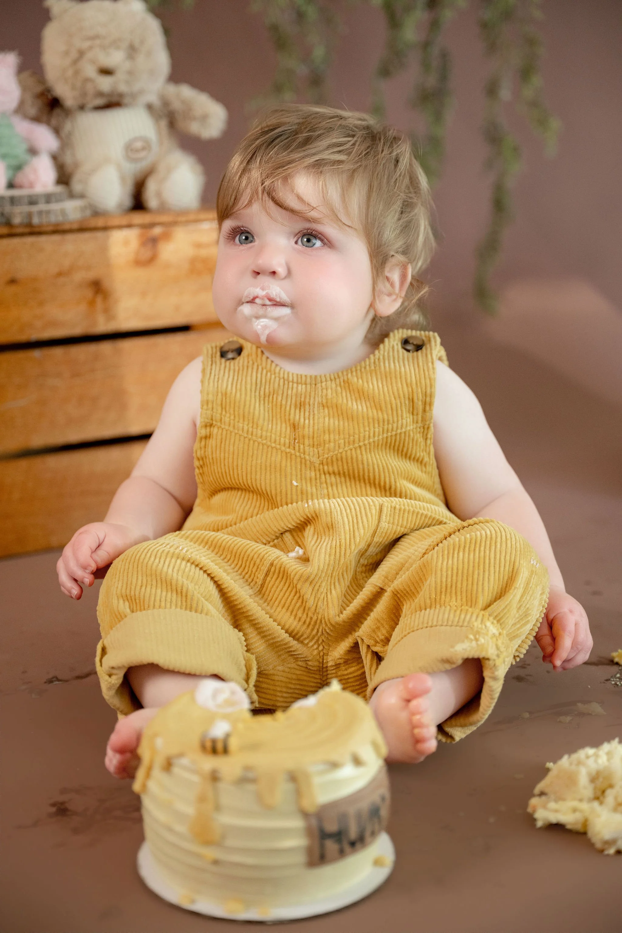 A young child with light brown hair and blue eyes, wearing a yellow corduroy overall, is sitting on the floor with a piece of cake in front of them. The child has frosting on their lips and face. In the background, a wooden box with a teddy bear and 
