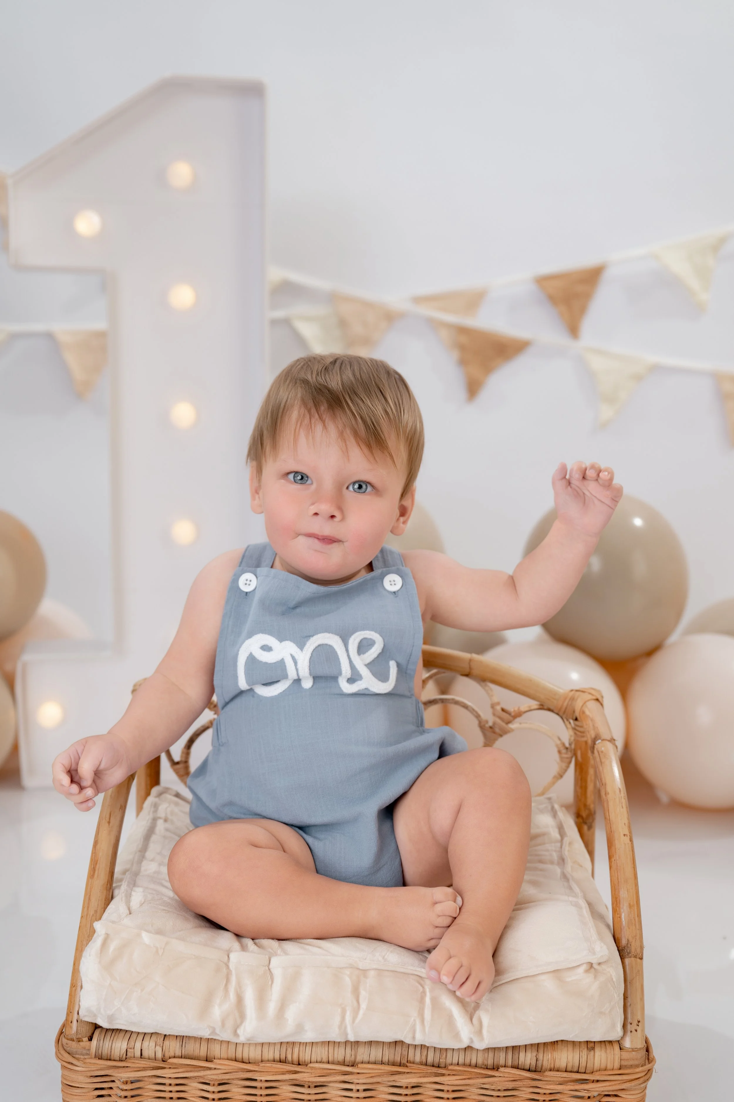 A young child with blue eyes and light brown hair sitting in a wicker chair, wearing a gray sleeveless romper with the word 'one' embroidered on it, against a neutral background decorated with balloons, bunting, and illuminated marquee numbers.
