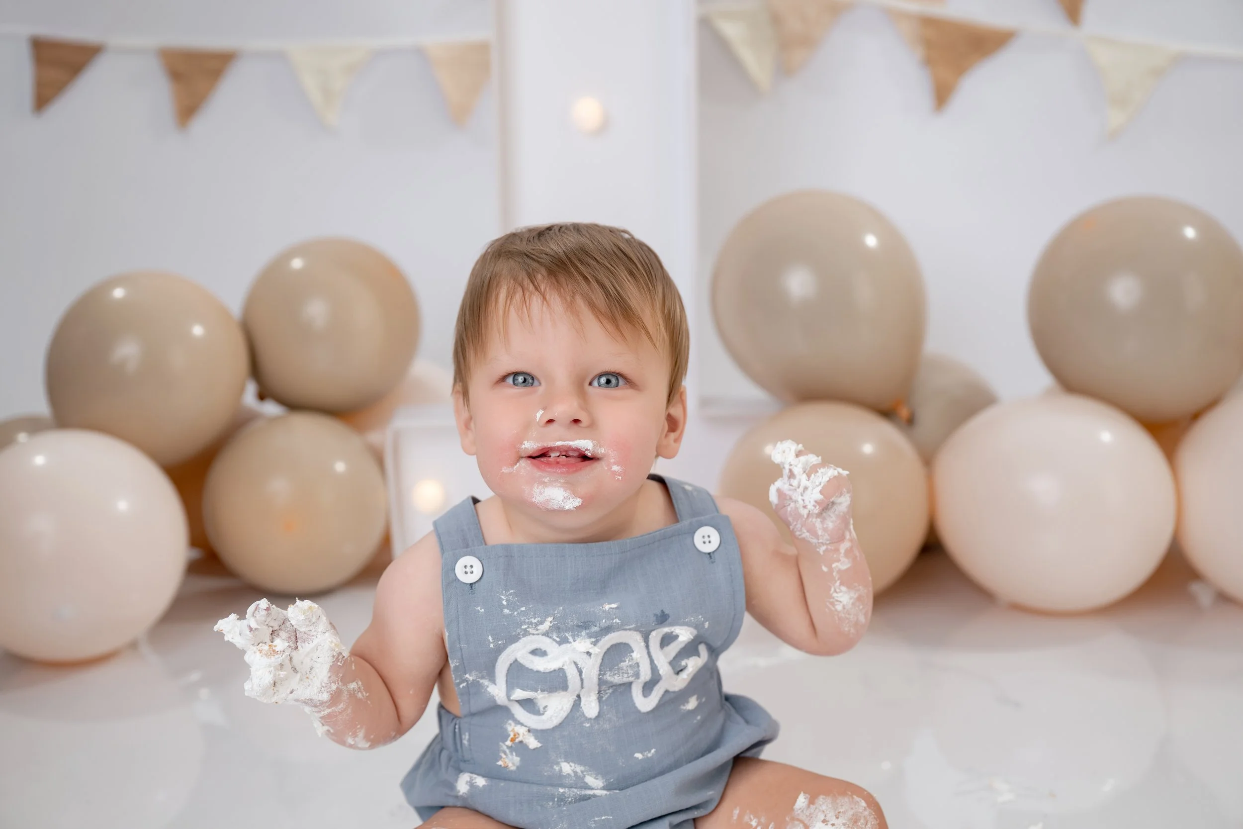 A young child with blue eyes and short light brown hair smiling while covered in white cake or frosting, sitting in front of a backdrop decorated with beige and cream-colored balloons and a bunting banner.