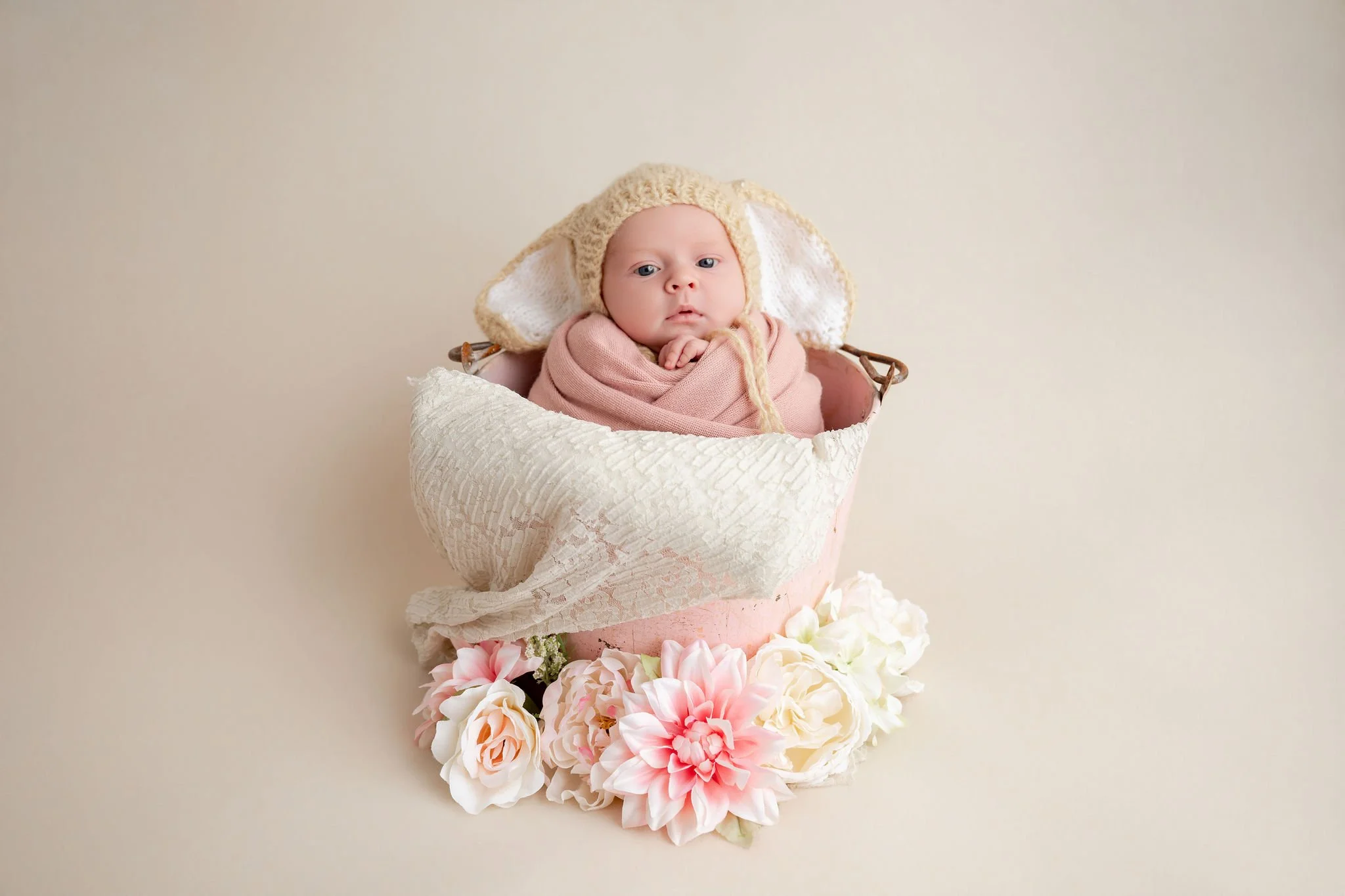 A baby dressed in pink wrapped in a pink blanket, wearing a knitted bonnet with floppy ears, sitting in a basket decorated with paper flowers.