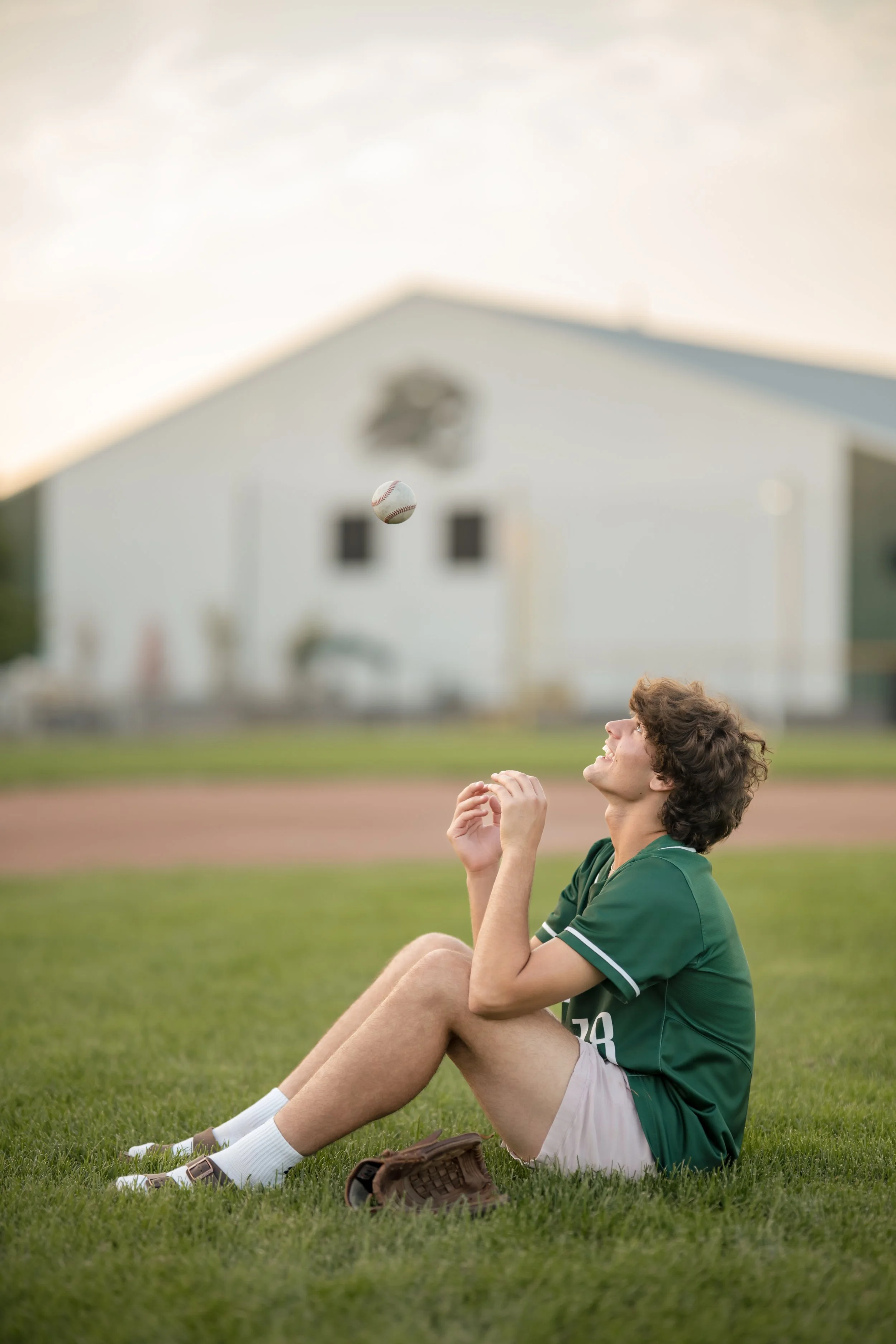 A young male baseball player with curly brown hair and light skin sitting on grass on a baseball field, smiling and looking up at a baseball in mid-air, with a baseball glove near his feet and a school building in the background during sunset.