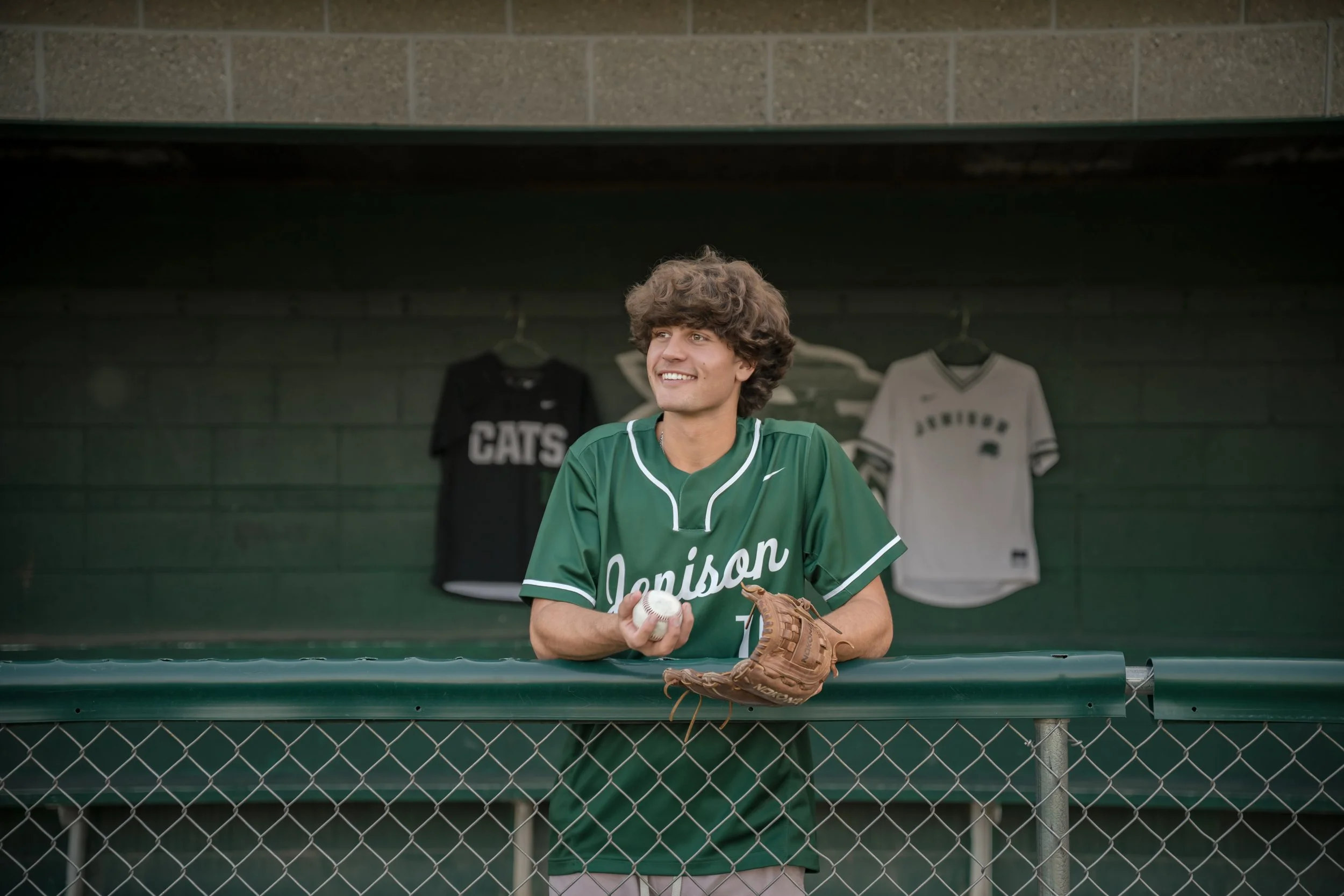 A young man wearing a green baseball jersey with 'Denison' written on it, holding a baseball and wearing a baseball glove, standing behind a green fence in a dugout with baseball jerseys hanging in the background.