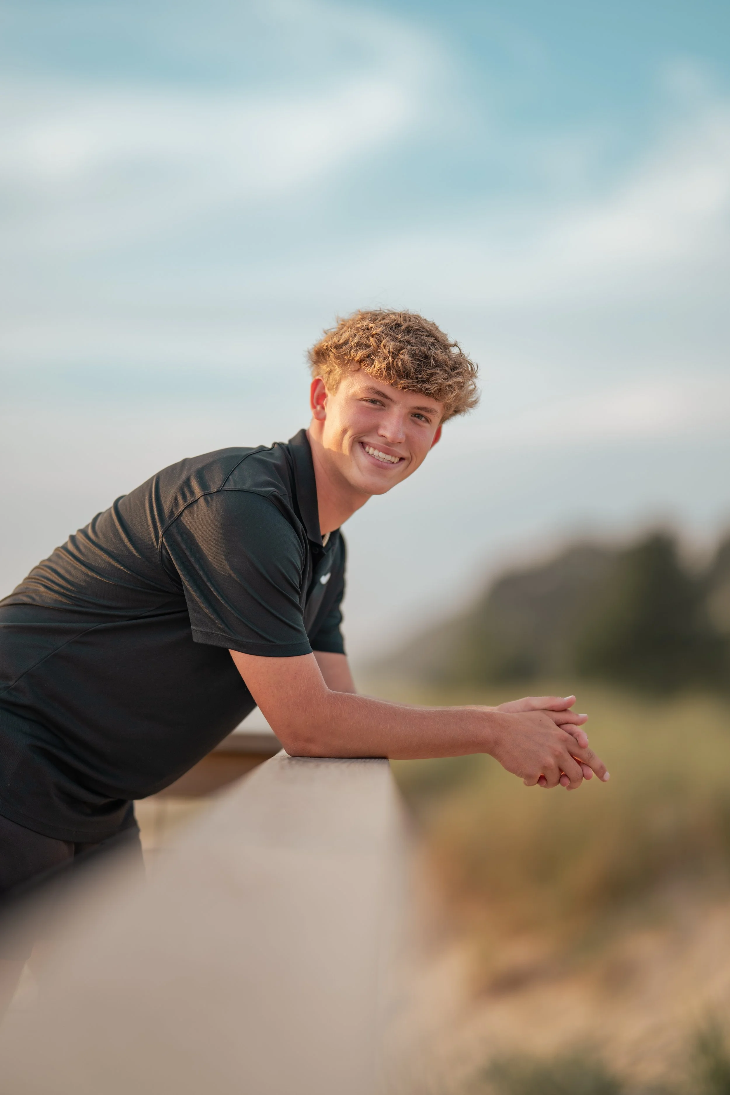 A young man with curly blond hair smiling and leaning on a wooden railing outdoors during daytime.