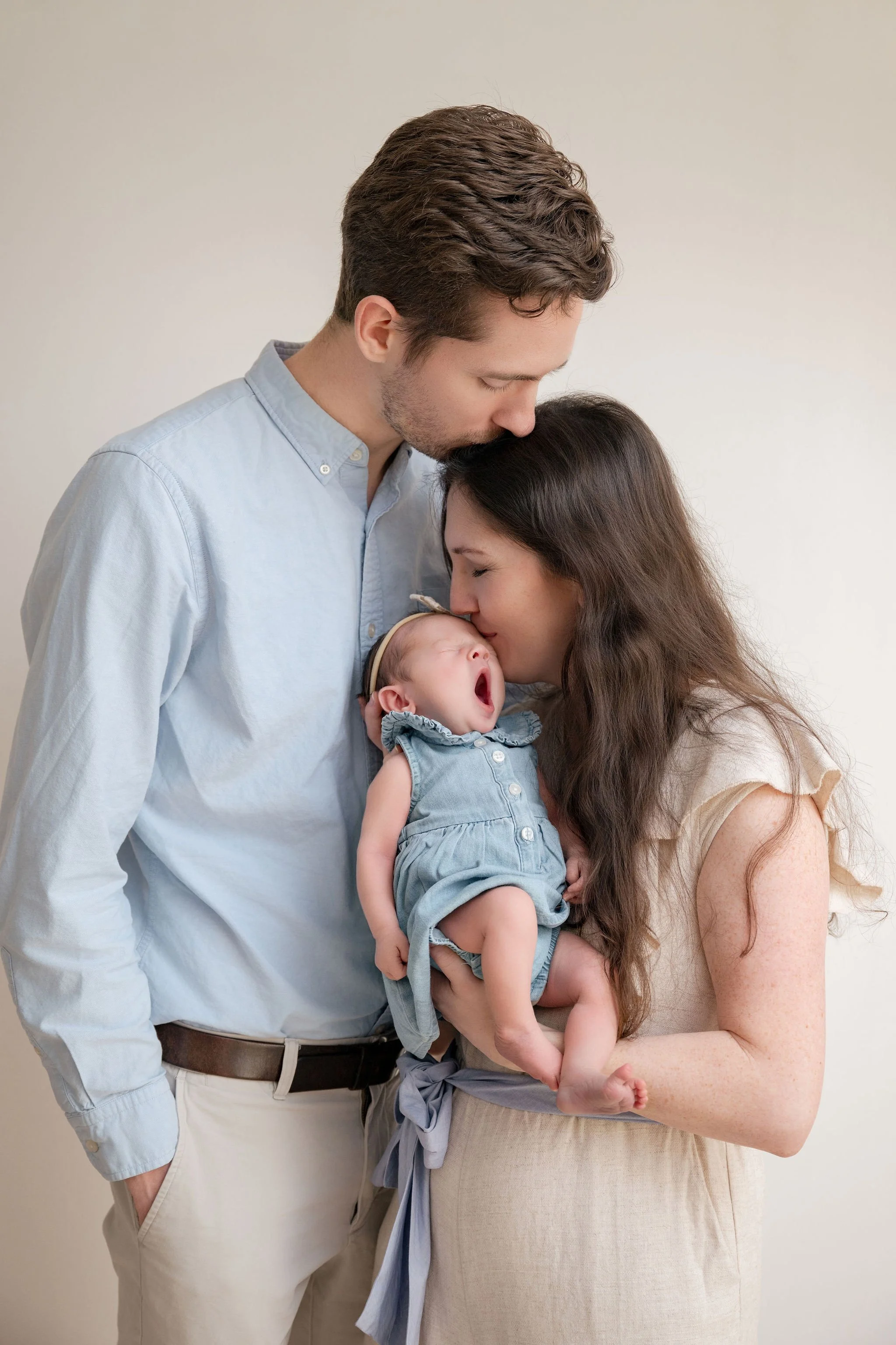 A family moment with a man, woman, and a baby. The man has brown hair, a pale blue shirt, and is kissing the baby's forehead. The woman has long, dark hair, and is holding the baby, who is yawning and dressed in a light denim outfit. The background is plain and off-white.