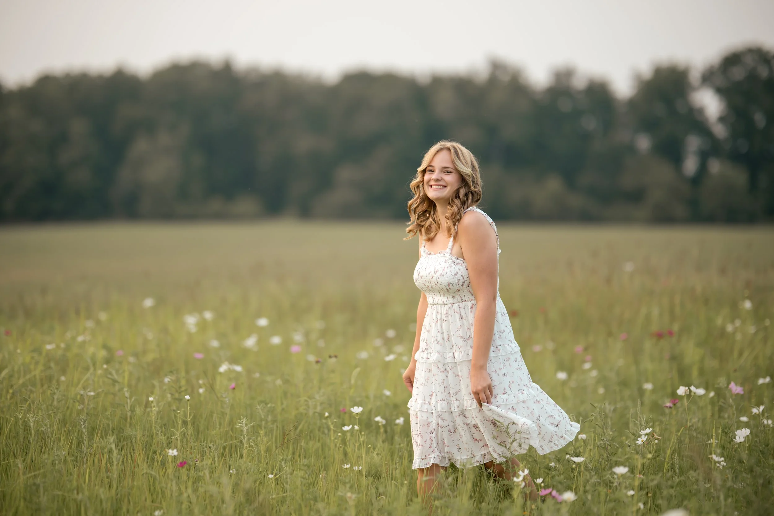 High school senior walking through a wildflower field wearing a white floral dress during golden hour. Muskegon senior photography