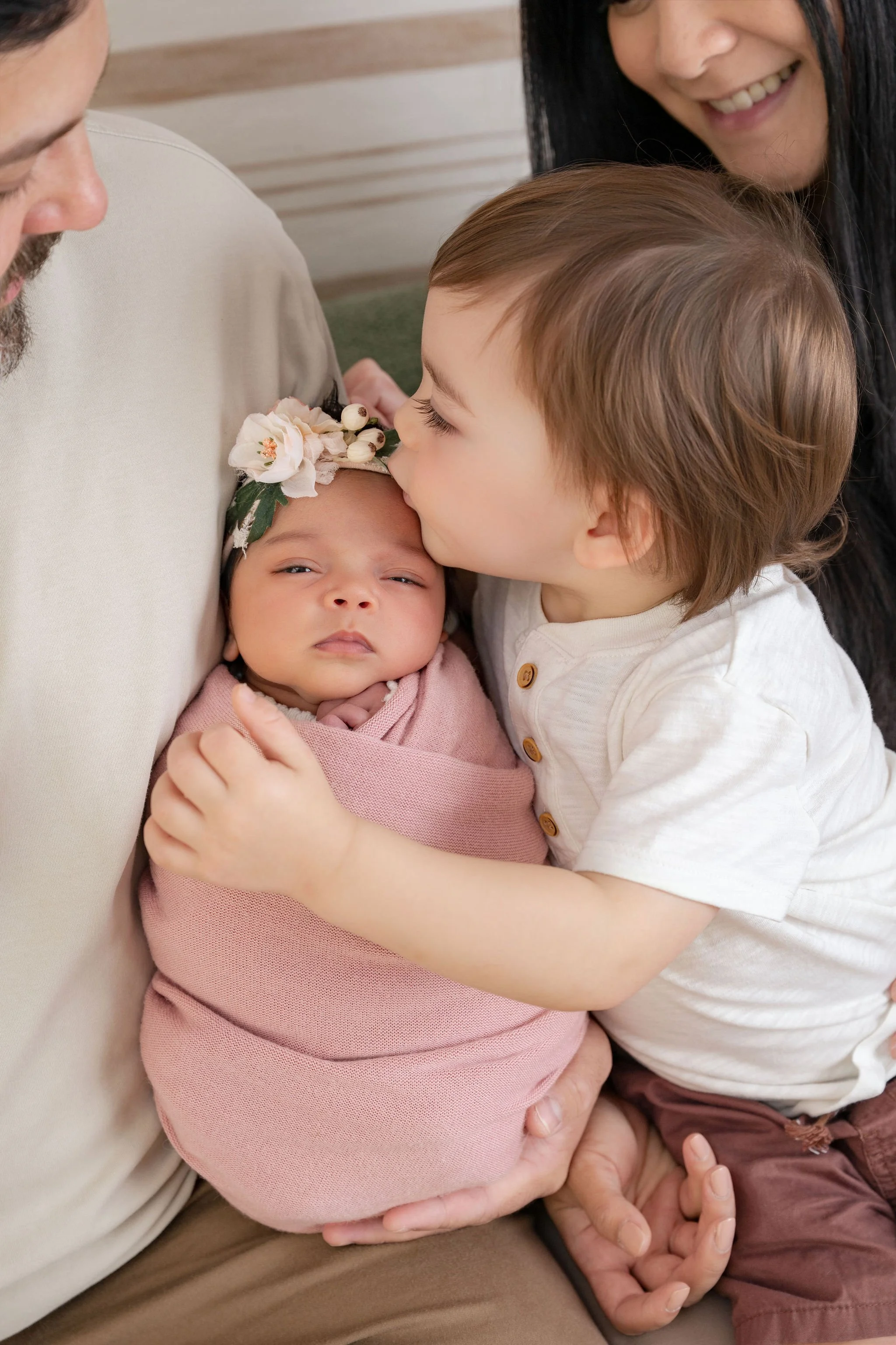A young child kissing a baby girl on the forehead while seated on an adult's lap, with another adult smiling nearby.