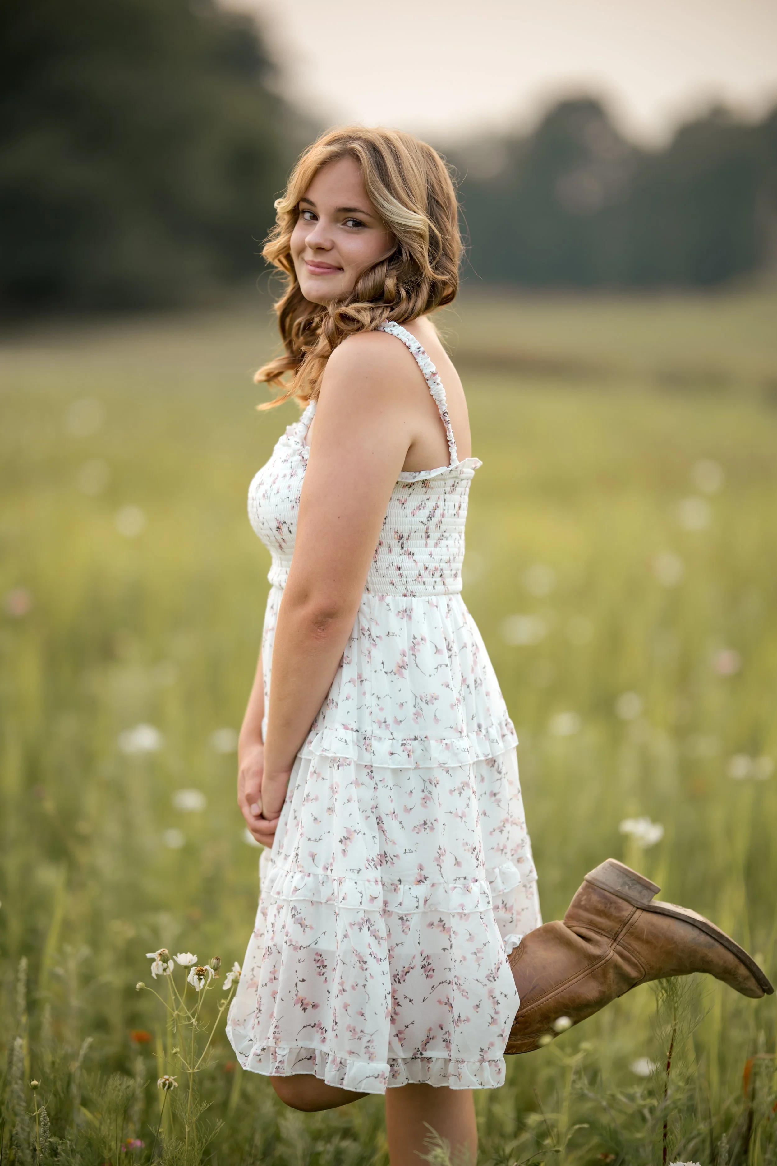 A young woman in a floral white summer dress kneeling in a field of flowers, with a gentle smile, tan boots, and curly hair, during sunset or golden hour.
