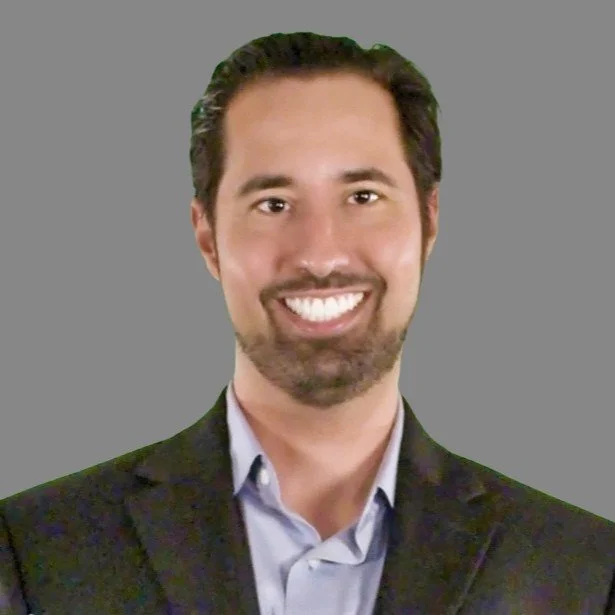 Smiling man with dark hair and beard wearing a dark blazer and light blue shirt, gray background.