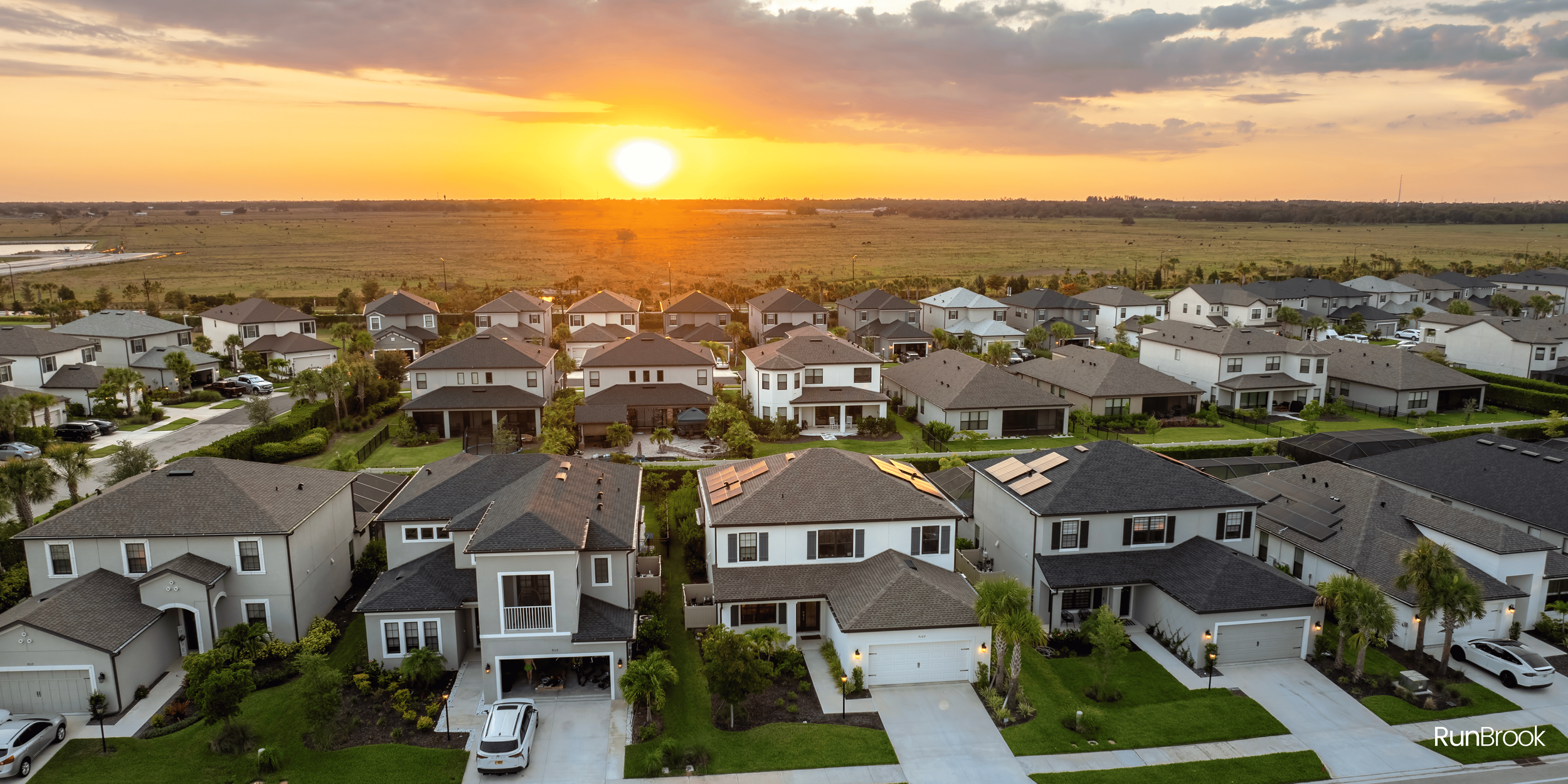Aerial view of a suburban neighborhood during sunset, showing houses with yards, driveways, and solar panels on some roofs near open fields.