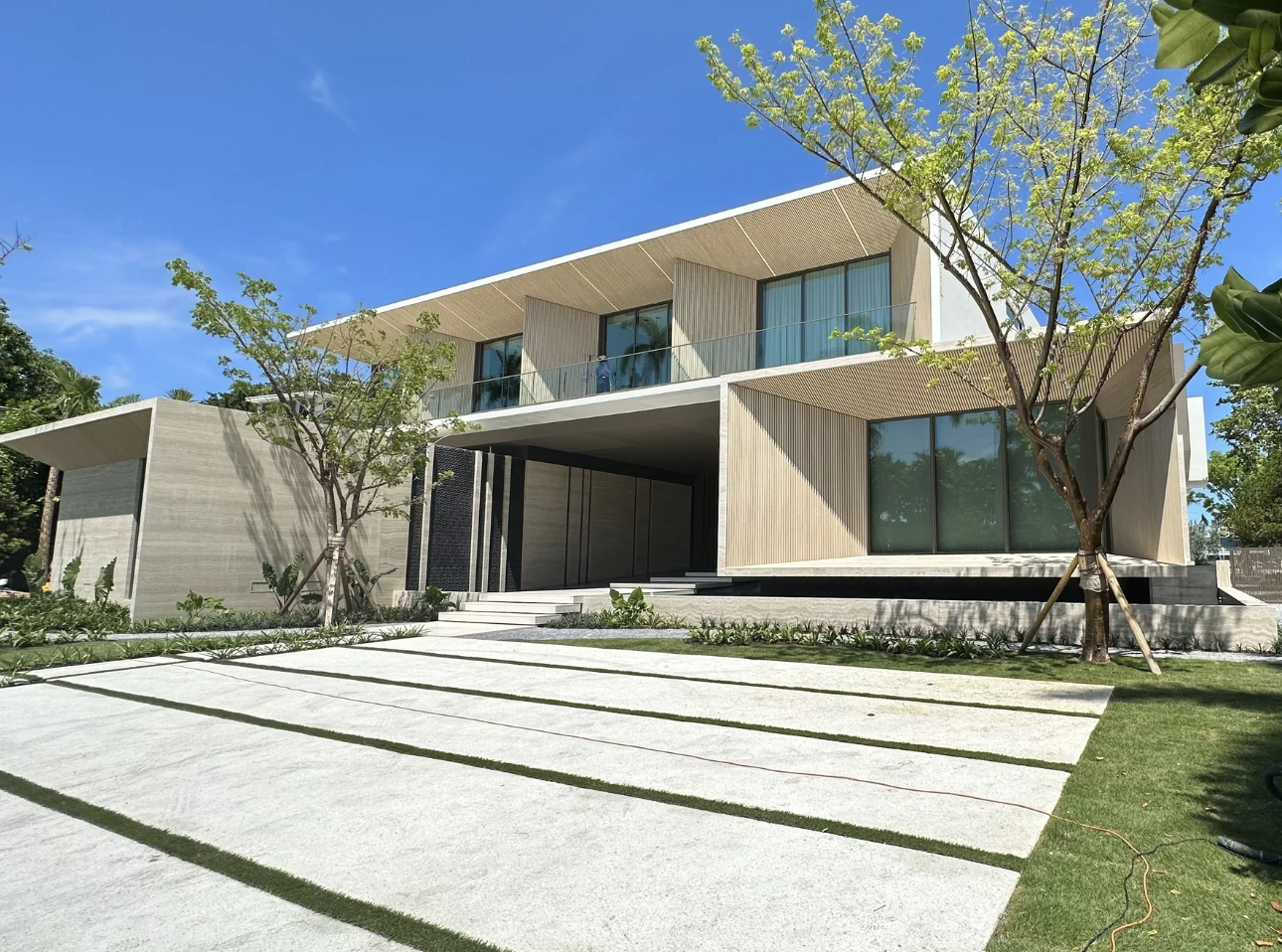 Modern two-story house with large glass windows, wooden accents, and a driveway surrounded by green grass and trees under a blue sky.