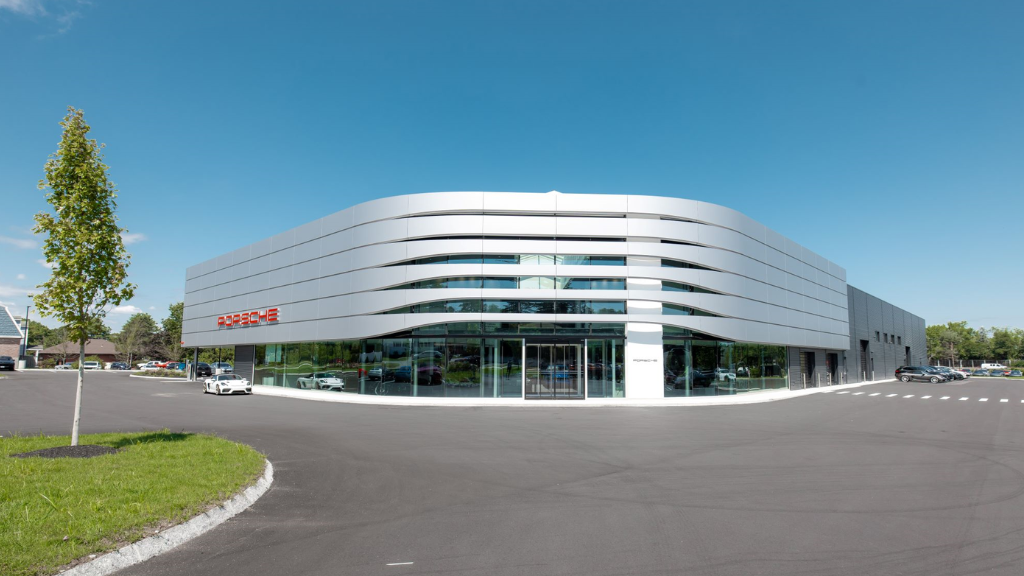 Modern car dealership building with Porsche logo, glass windows, and parking lot under a clear blue sky.