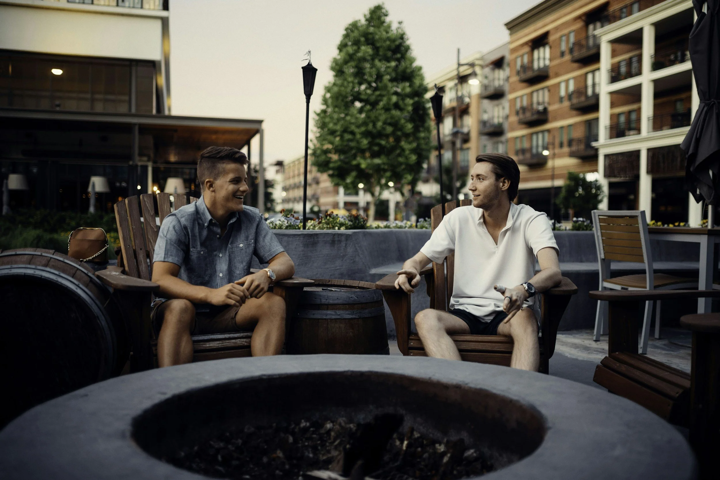 Two young men are sitting outdoors at a patio table, engaged in conversation and smiling. The scene is during the evening with a background of modern apartment buildings, trees, and patio decor, including an umbrella and string lights.