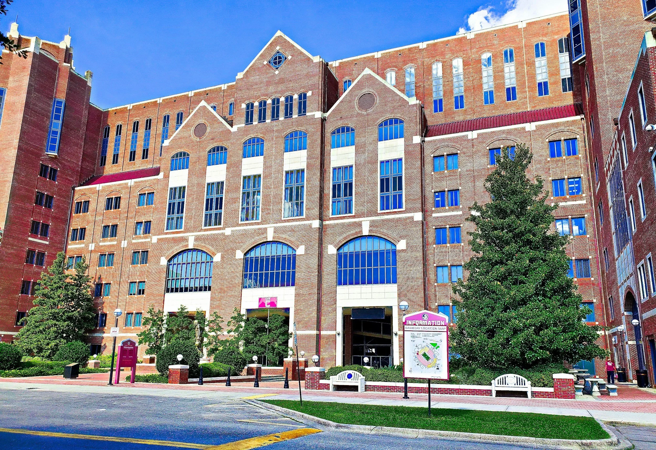 Red-brick multi-story building with large arched and rectangular windows, trees in front, a street, benches, and informational signage outside, under a blue sky with some clouds.