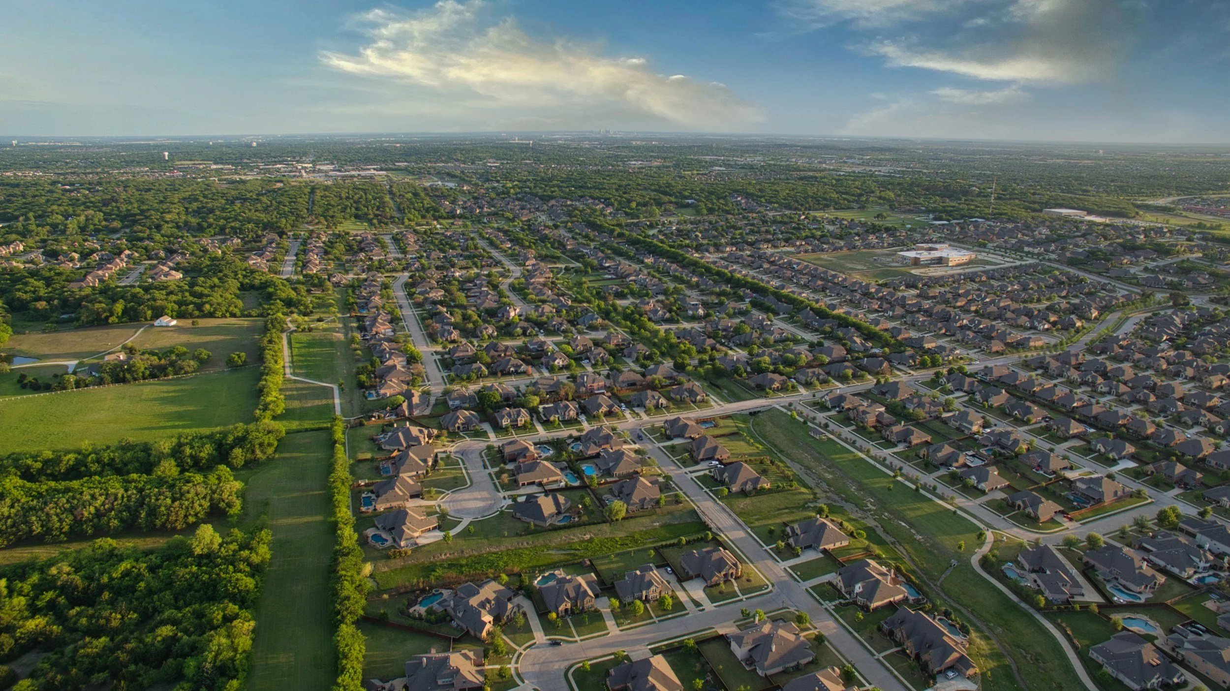 An aerial view of a suburban neighborhood with houses, streets, and green spaces, with a large park or wooded area on the left and a school or community center on the right, under a partly cloudy sky.