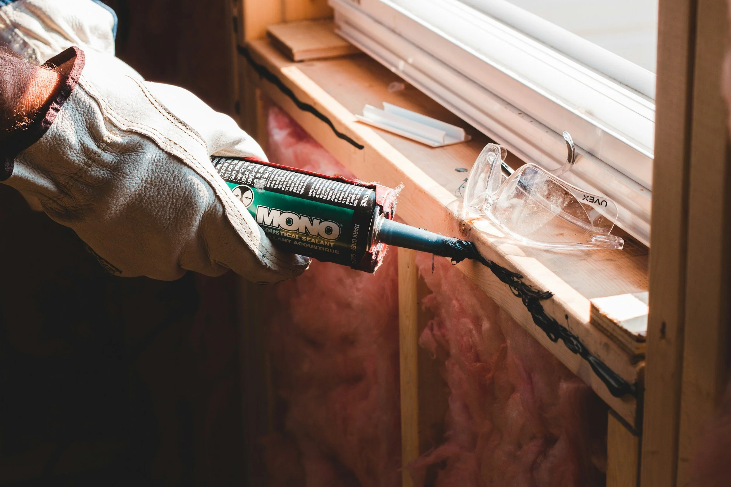Person working with caulking gun on wooden window frame in a room under construction.