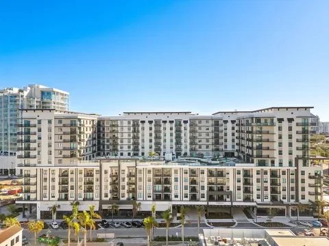 Modern multi-story apartment complex with palm trees and parking lot in foreground under clear blue sky.