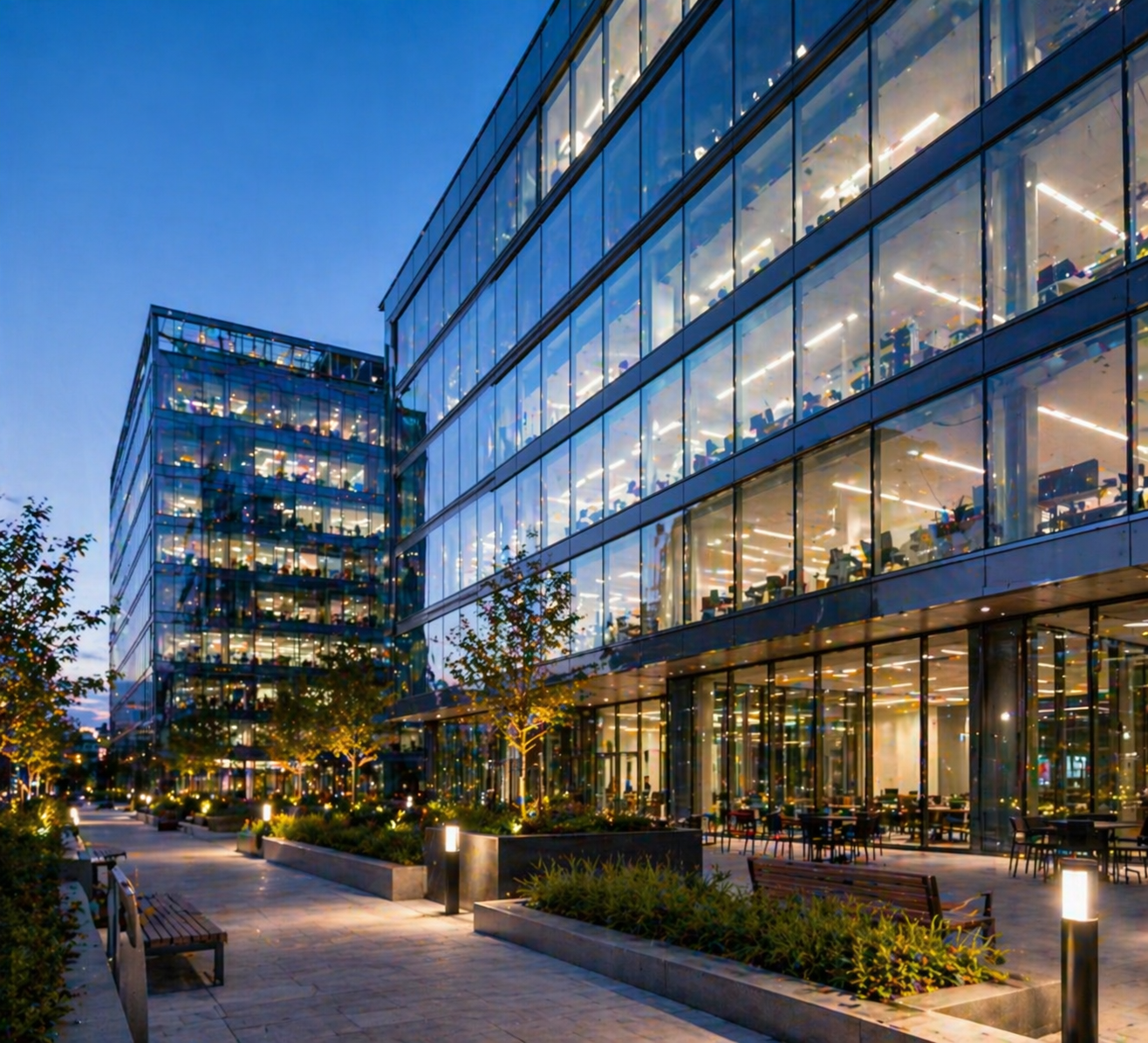 Exterior view of a modern glass office building at dusk, with illuminated interior spaces and outdoor seating areas surrounded by trees and pathways.