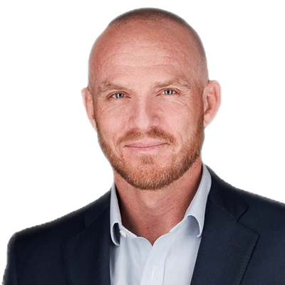 Close-up of a middle-aged man with a shaved head, light beard, wearing a navy blazer and white shirt, smiling against a plain white background.