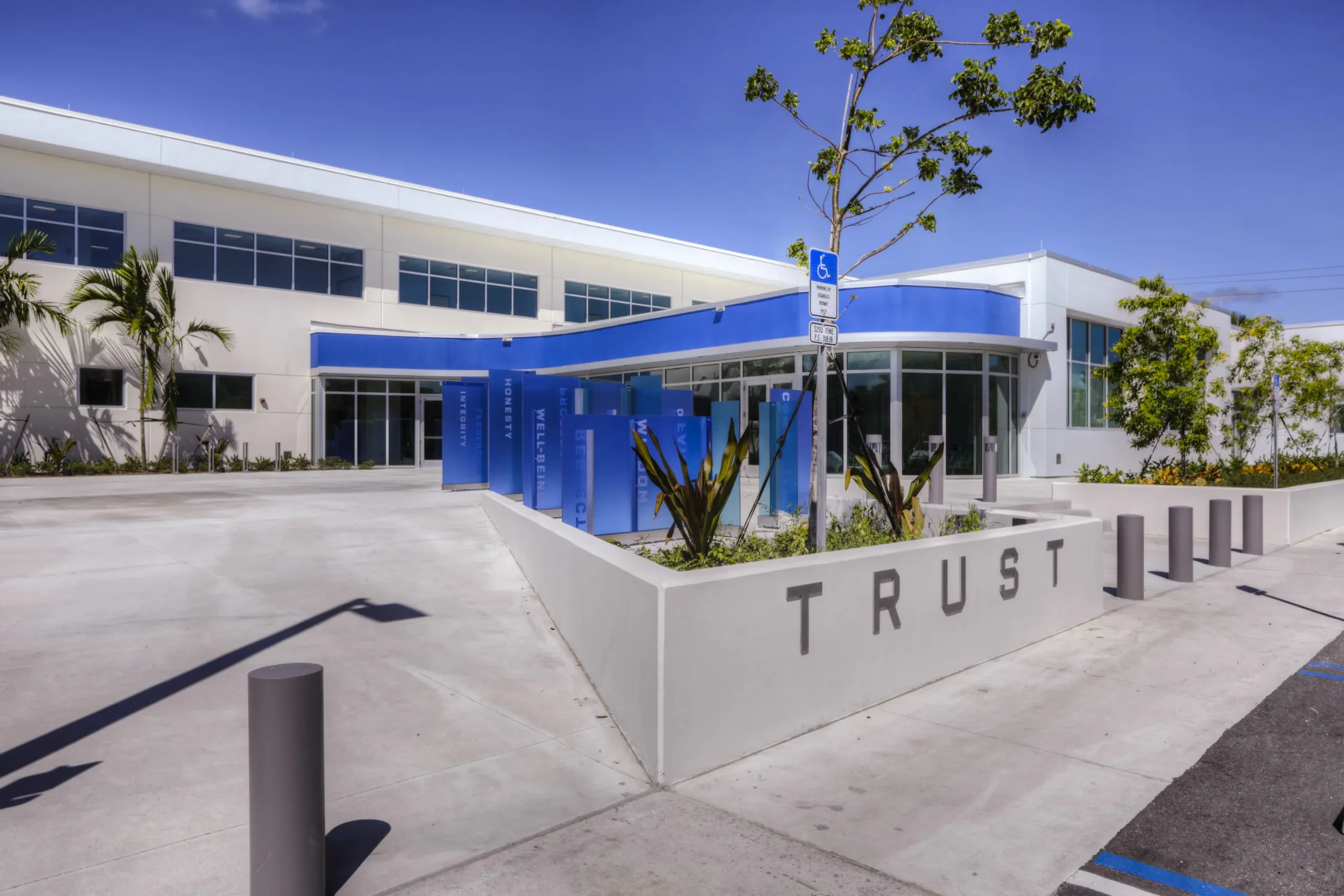 Modern building with glass windows, blue and white exterior, surrounded by landscaped plants, with a sidewalk and parking area in the foreground under a clear blue sky.